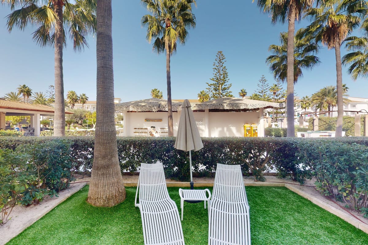 A well-maintained outdoor seating area with two white lounge chairs and a single umbrella, surrounded by lush greenery and palm trees.