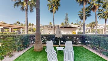 A well-maintained outdoor seating area with two white lounge chairs and a single umbrella, surrounded by lush greenery and palm trees.