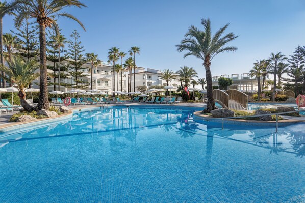 Una piscina con sillones de descanso y sombrillas, rodeada de palmeras y un edificio con balcones.