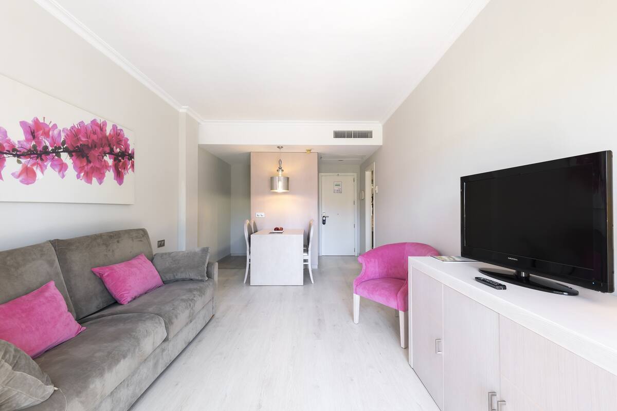 A modern living room with a grey sofa, a pink armchair, a flat-screen TV, and a dining area with a white table and chairs.