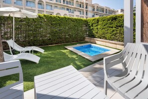 Un área de piscina con sillas reclinables blancas y una sombrilla, rodeada de césped y setos cuidados, con un edificio al fondo.