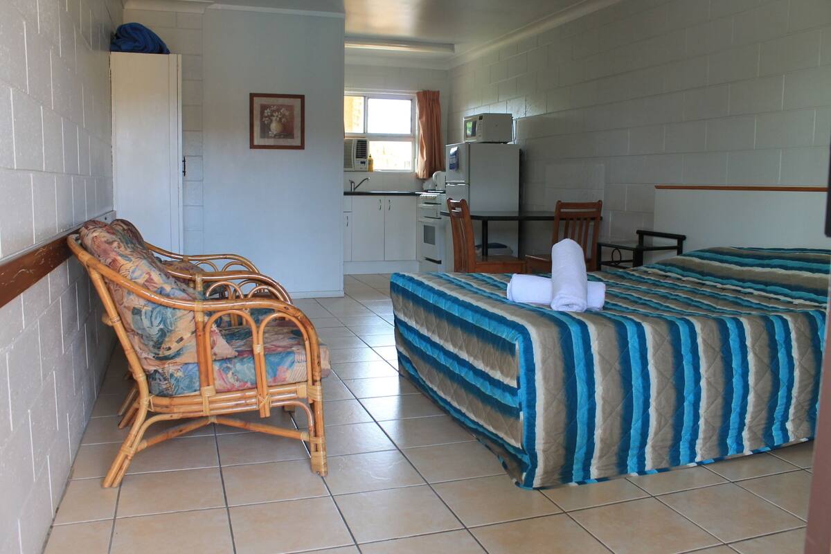 A room with a bed, a chair, a desk, a microwave, and a window.
