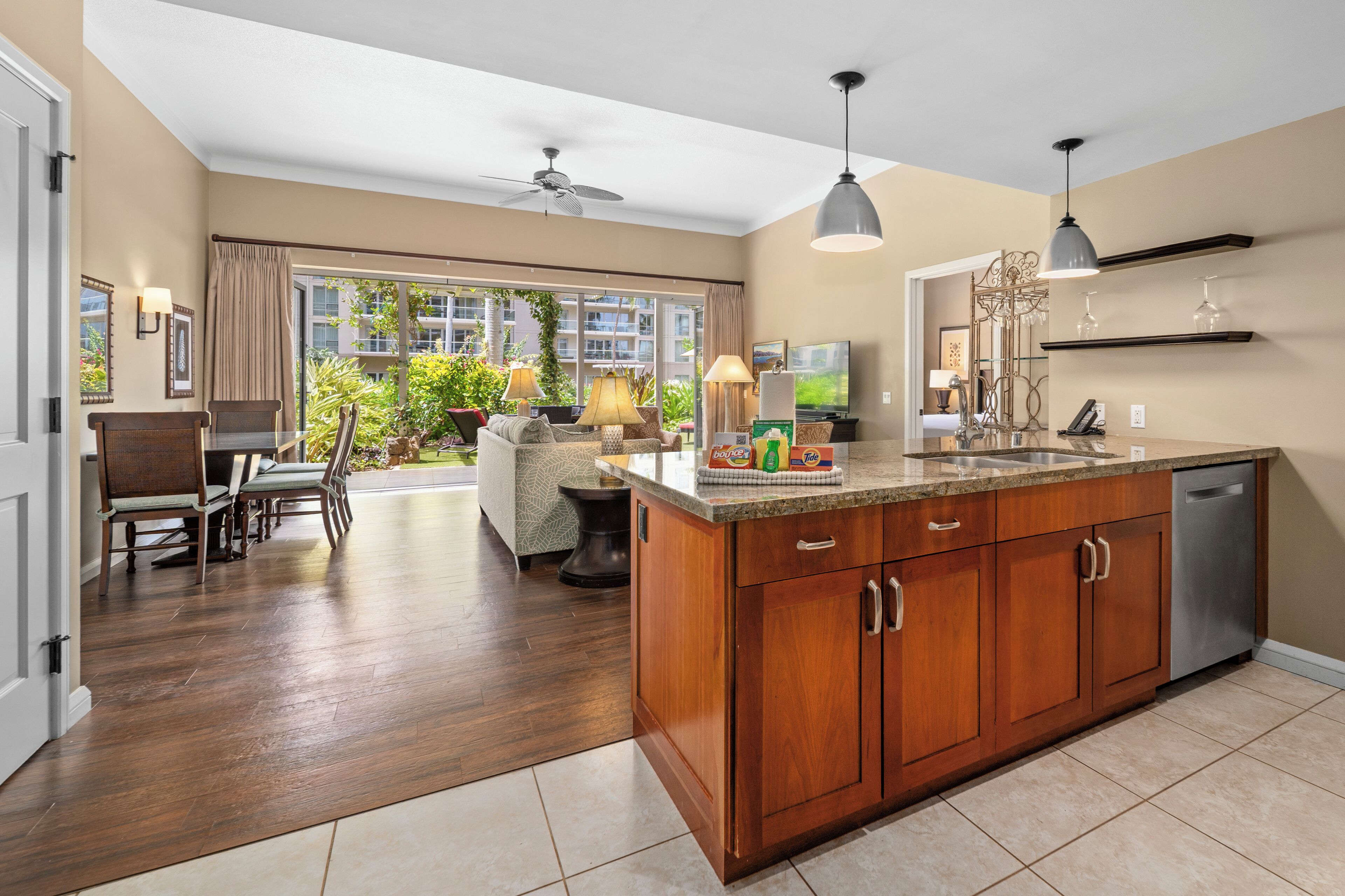 A spacious kitchen with a large island, wooden cabinets, and a dishwasher. There is a dining area with a table and chairs, and a view of a balcony with plants.