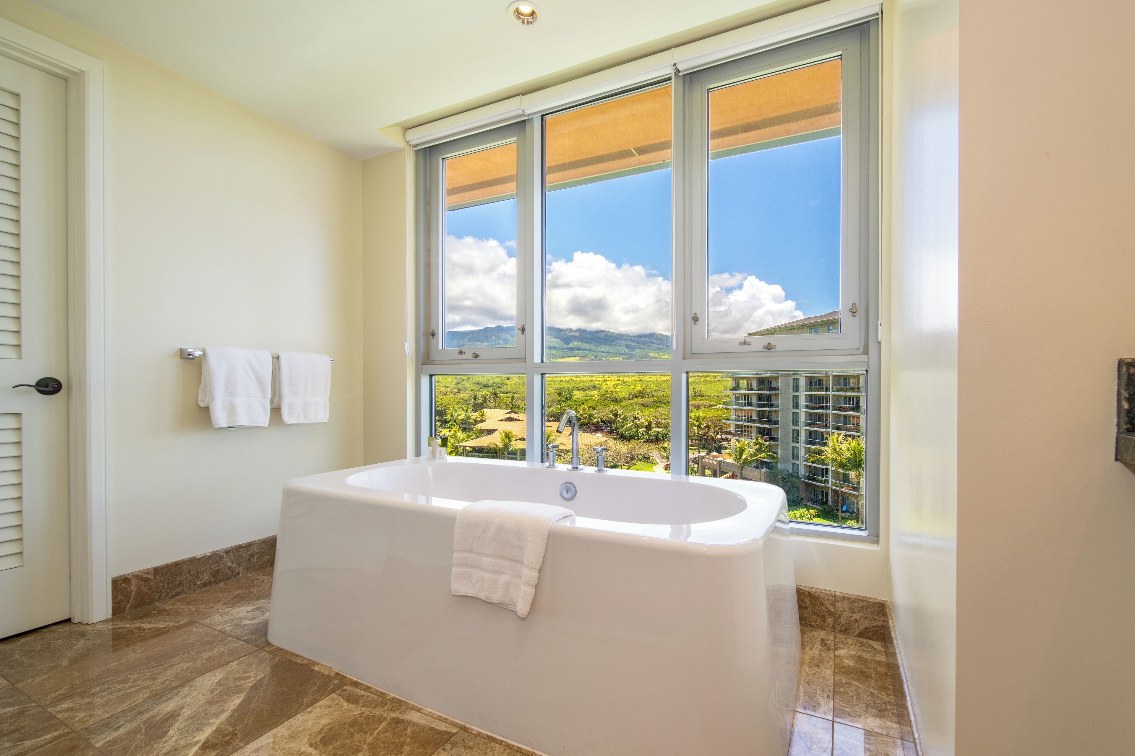A bathroom with a large bathtub, a window with a view of greenery and buildings, and a door with a towel hanging on the wall.