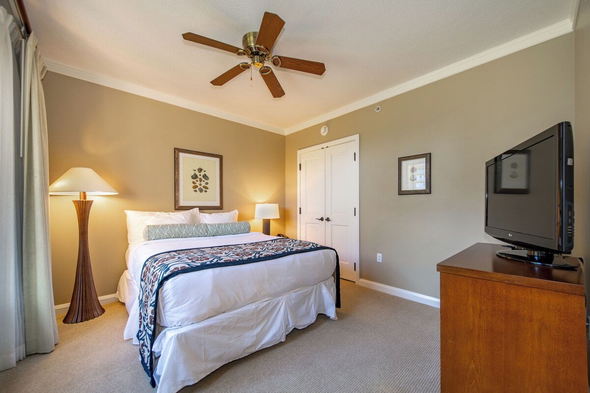 A hotel room with a bed, a ceiling fan, a TV, and a door.