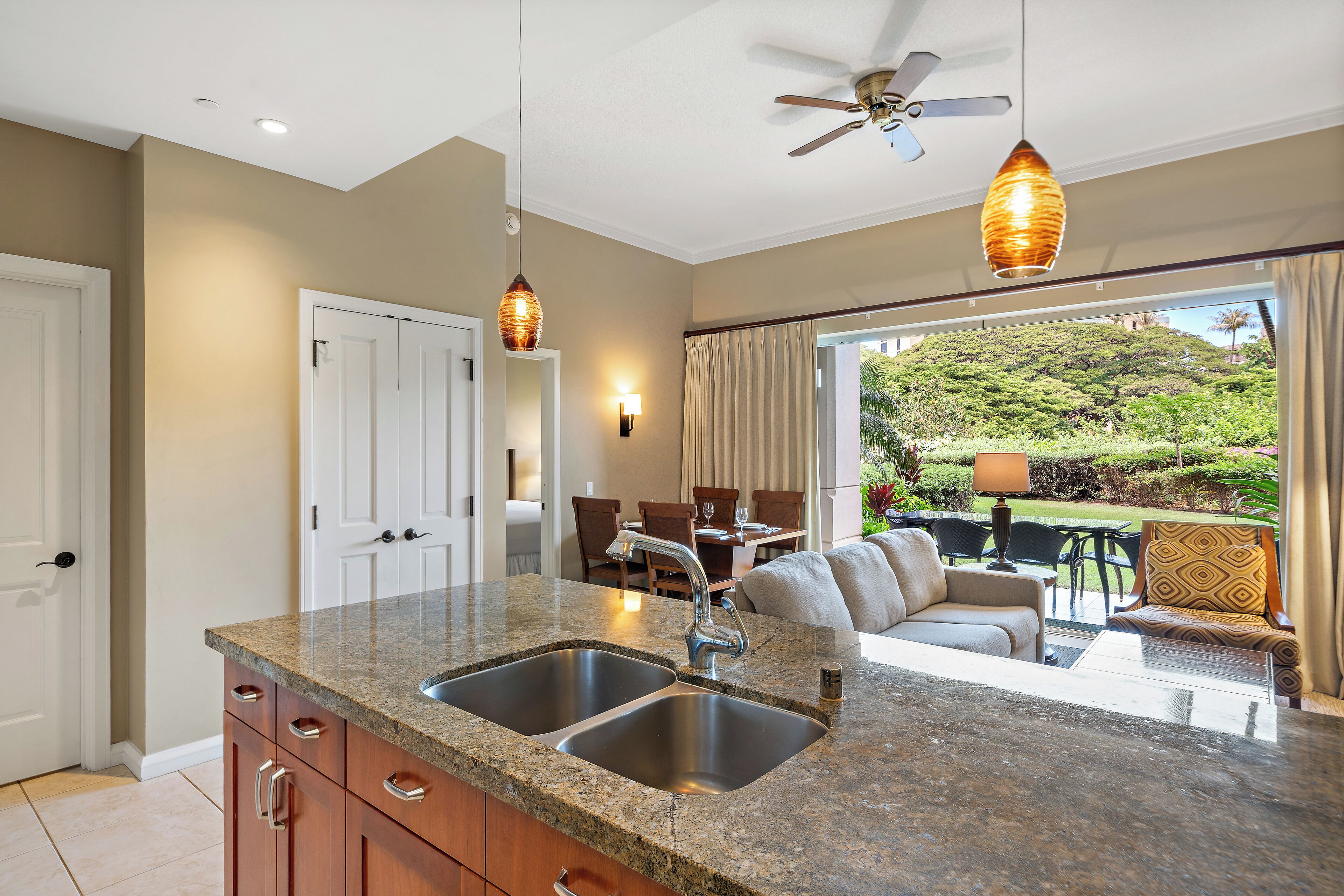 A kitchen with granite countertops, stainless steel sinks, and wooden cabinets. There is a ceiling fan and pendant lights.
