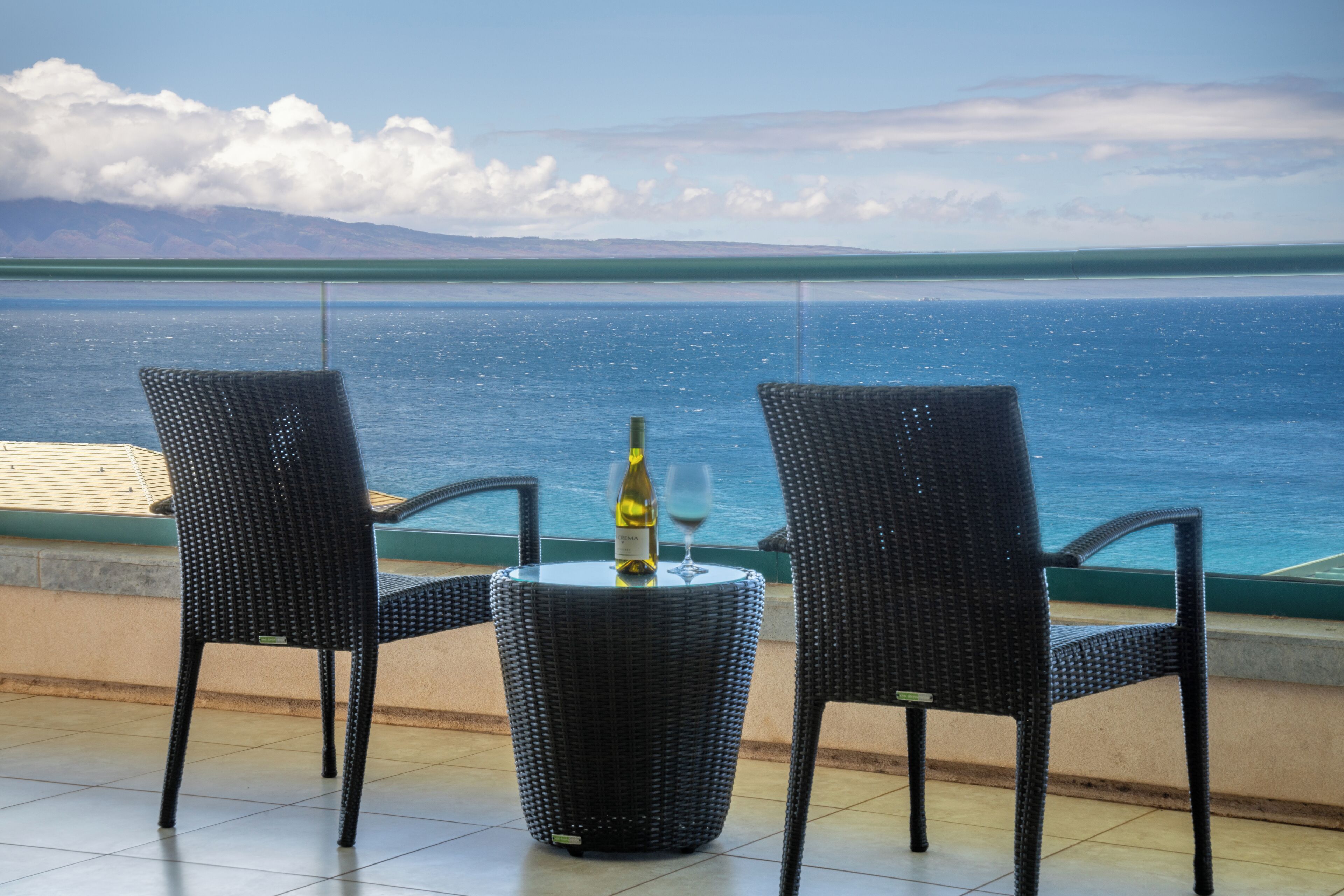 Een terras met twee rieten stoelen en een kleine tafel, een fles wijn en een glas op de tafel. Het terras kijkt uit over een waterpartij met bergen op de achtergrond.