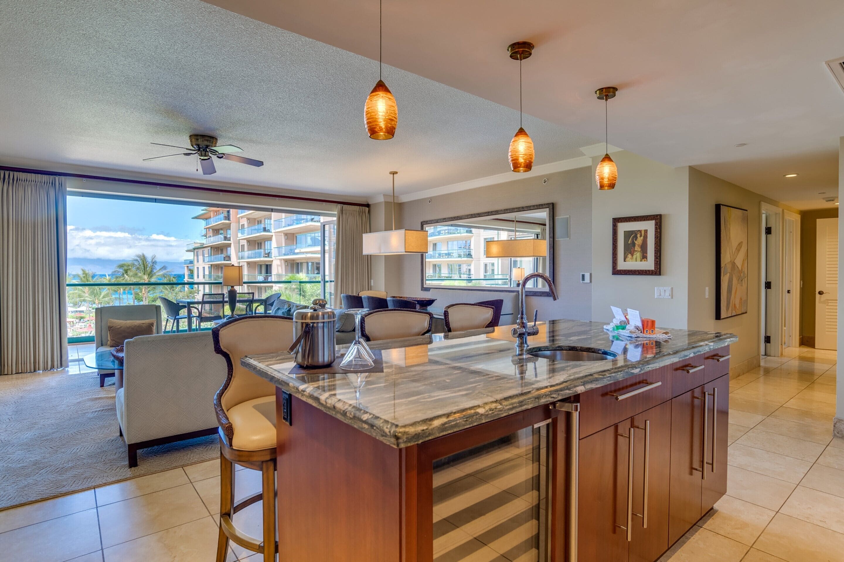 Une cuisine moderne dotée d’un grand îlot central, de luminaires suspendus et offrant une vue sur un balcon aménagé avec du mobilier d’extérieur.