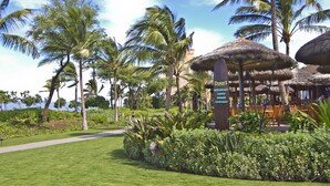 A tropical resort with thatched-roof structures, palm trees, and a sign indicating breakfast, lunch, dinner, and cocktails. - OUTRIGGER Honua Kai Resort & Spa (Lahaina)