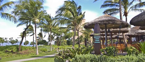 Un resort tropical con construcciones de techo de paja, palmeras y un cartel que indica desayuno, almuerzo, cena y cócteles.
