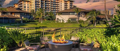 Um pátio com lareira e cadeiras, rodeado por vegetação exuberante e um edifício de hotel ao fundo.