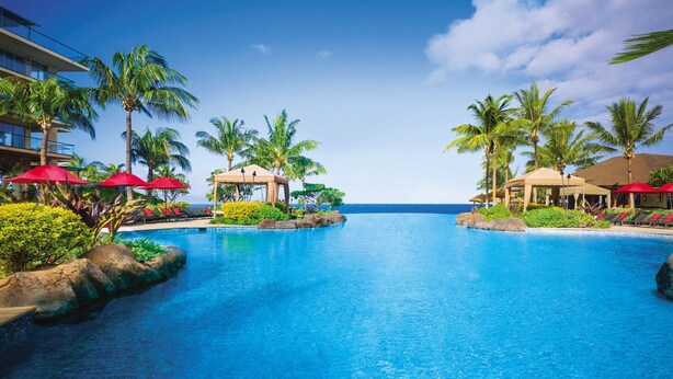 Une piscine d’hôtel avec des parasols rouges, des palmiers et une vue sur l’océan.