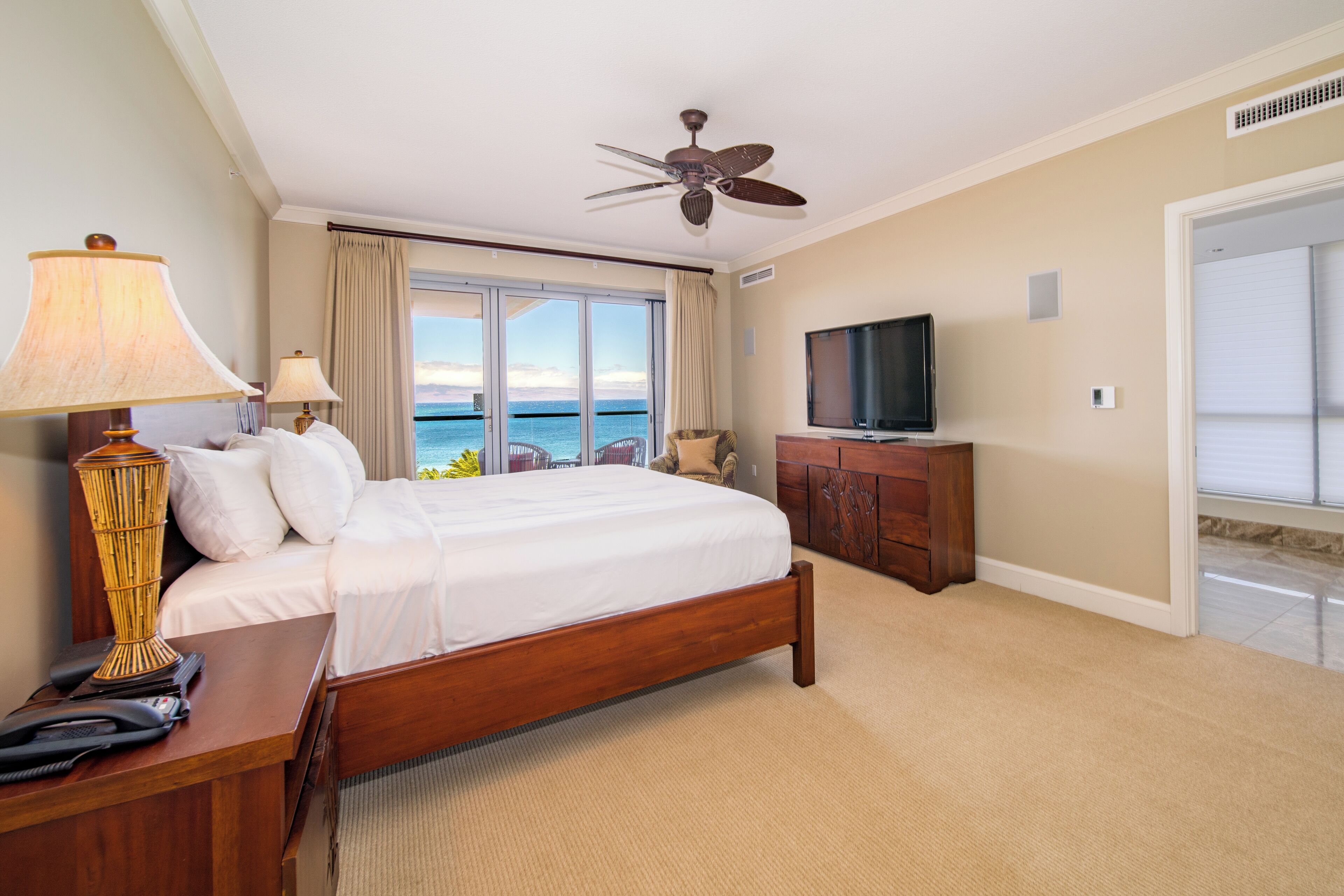 Habitación de hotel con cama, televisión, escritorio con lámpara, ventilador de techo y vista al mar.
