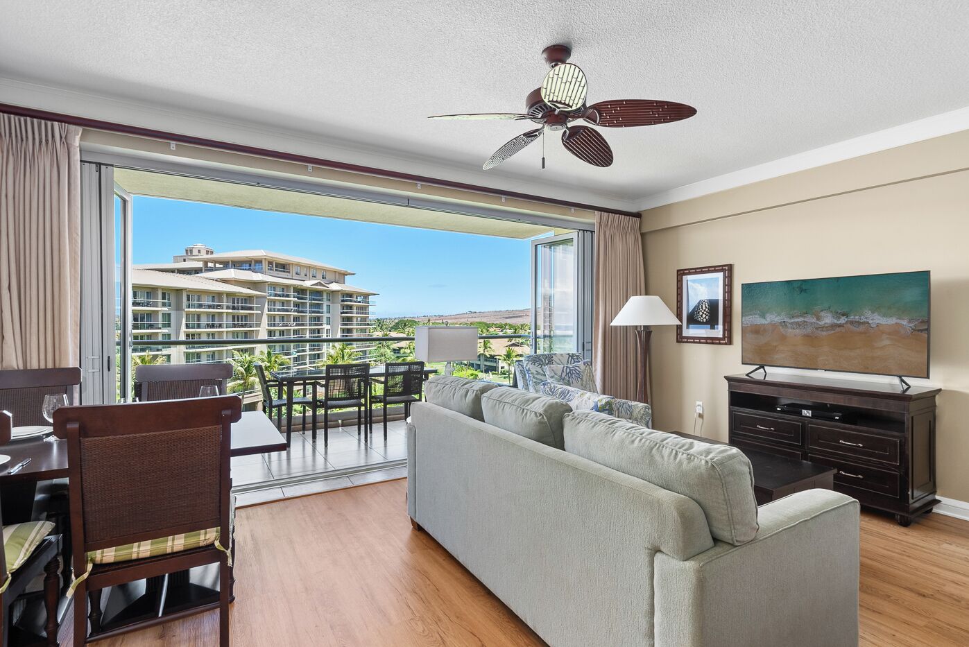 A hotel room with a balcony, a sofa, a dining table with chairs, a TV, and a ceiling fan.