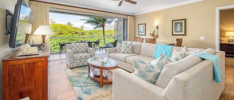 A spacious living room with a large sofa, a coffee table, and a television. There is a balcony with a view of greenery and a ceiling fan.