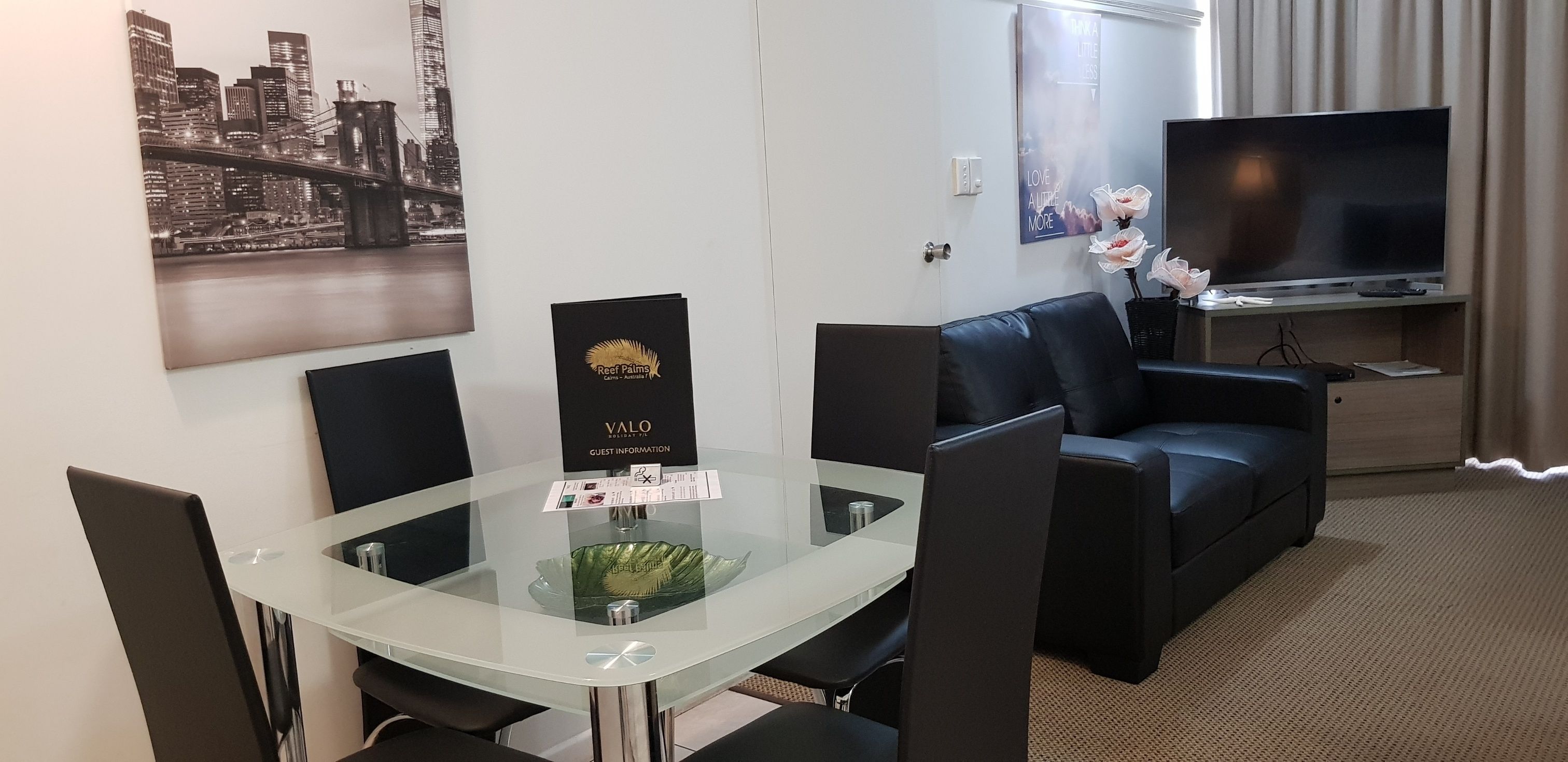 Hotelkamer met een glazen tafel, een zwart leren bank, een flatscreen televisie en een poster van een stadsgezicht.