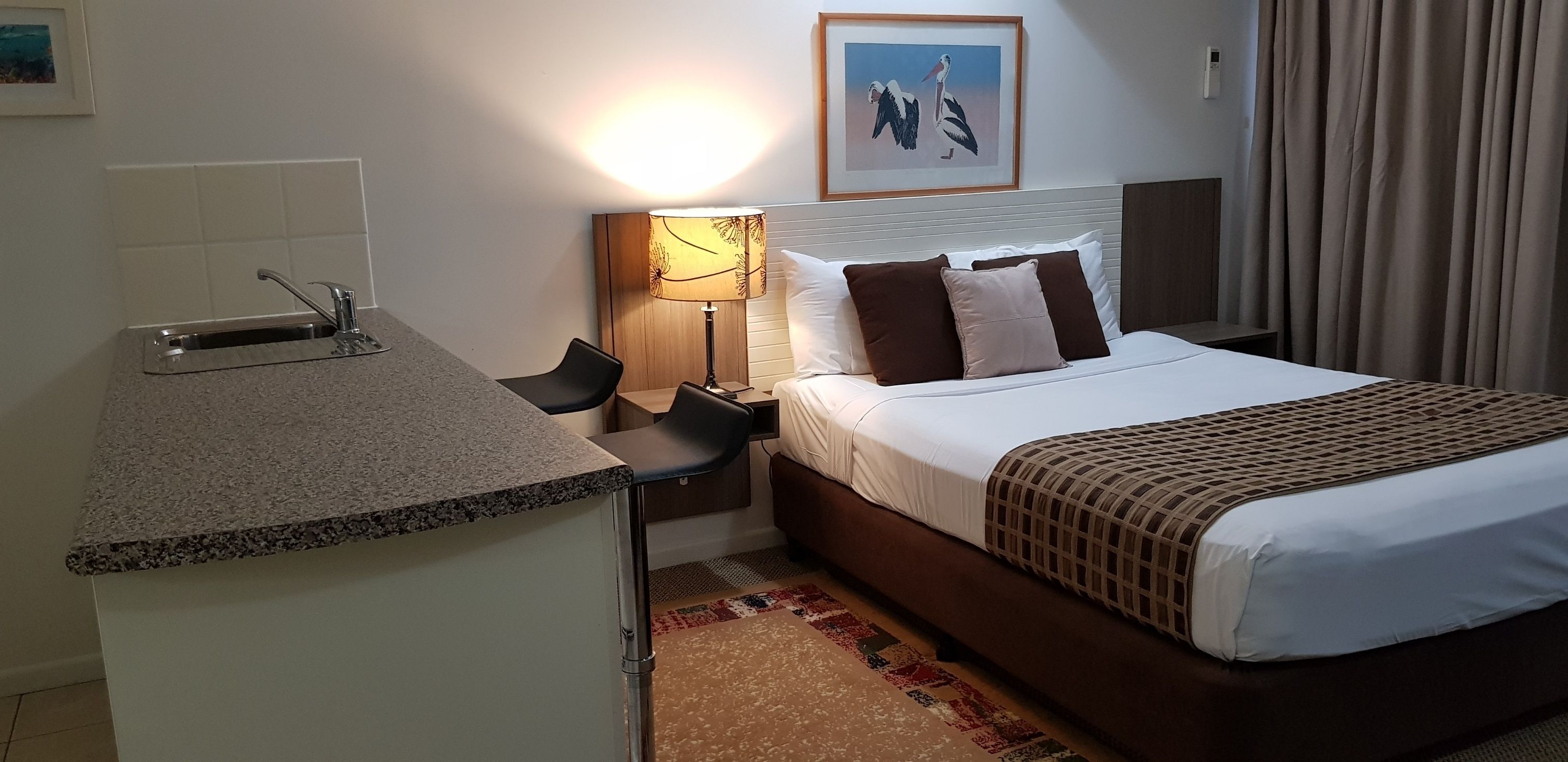 Ett hotellrum med en säng, ett skrivbord med handfat, en lampa och en inramad tavla på väggen.