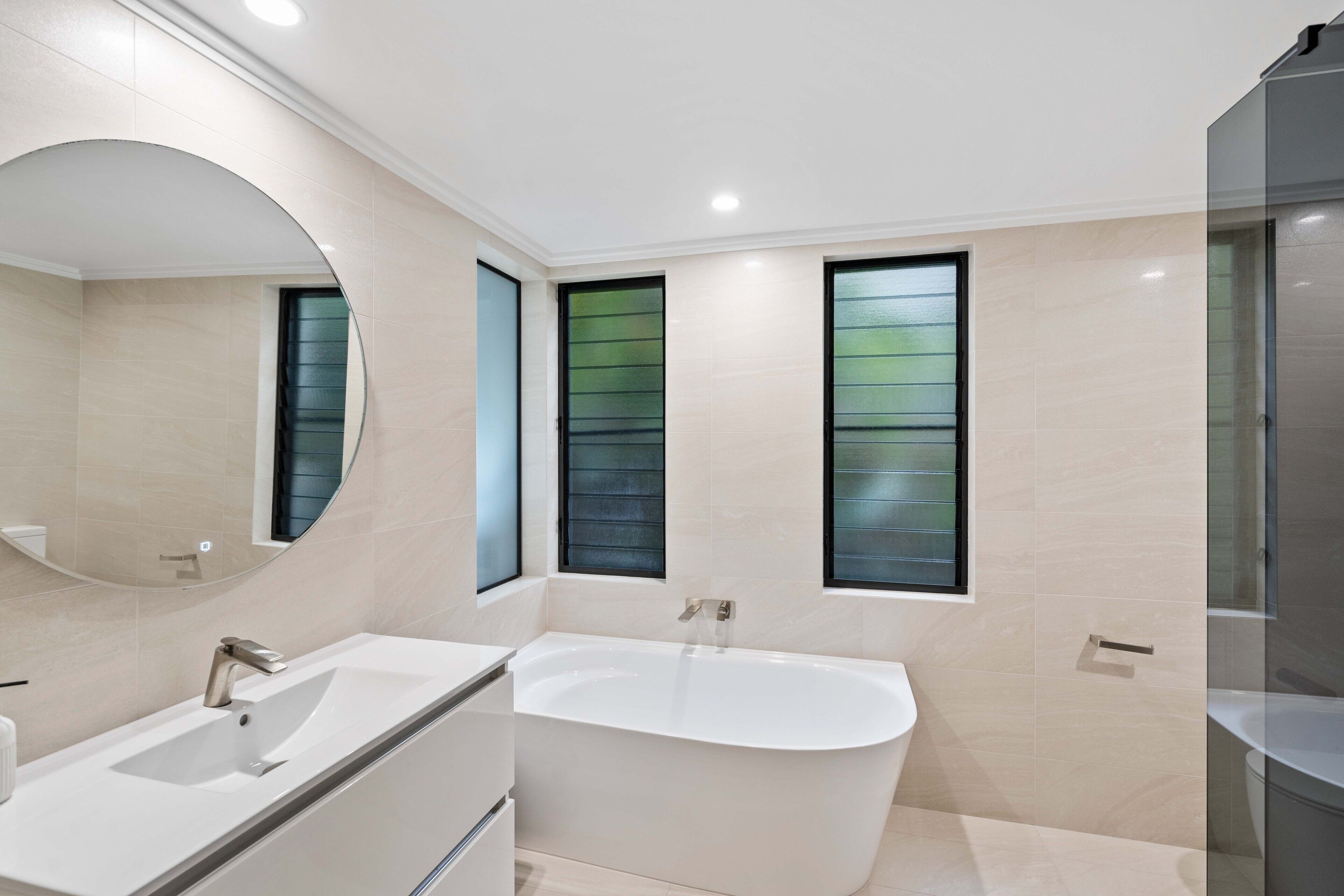 A modern bathroom with a freestanding bathtub, a large mirror, and two windows with frosted glass.