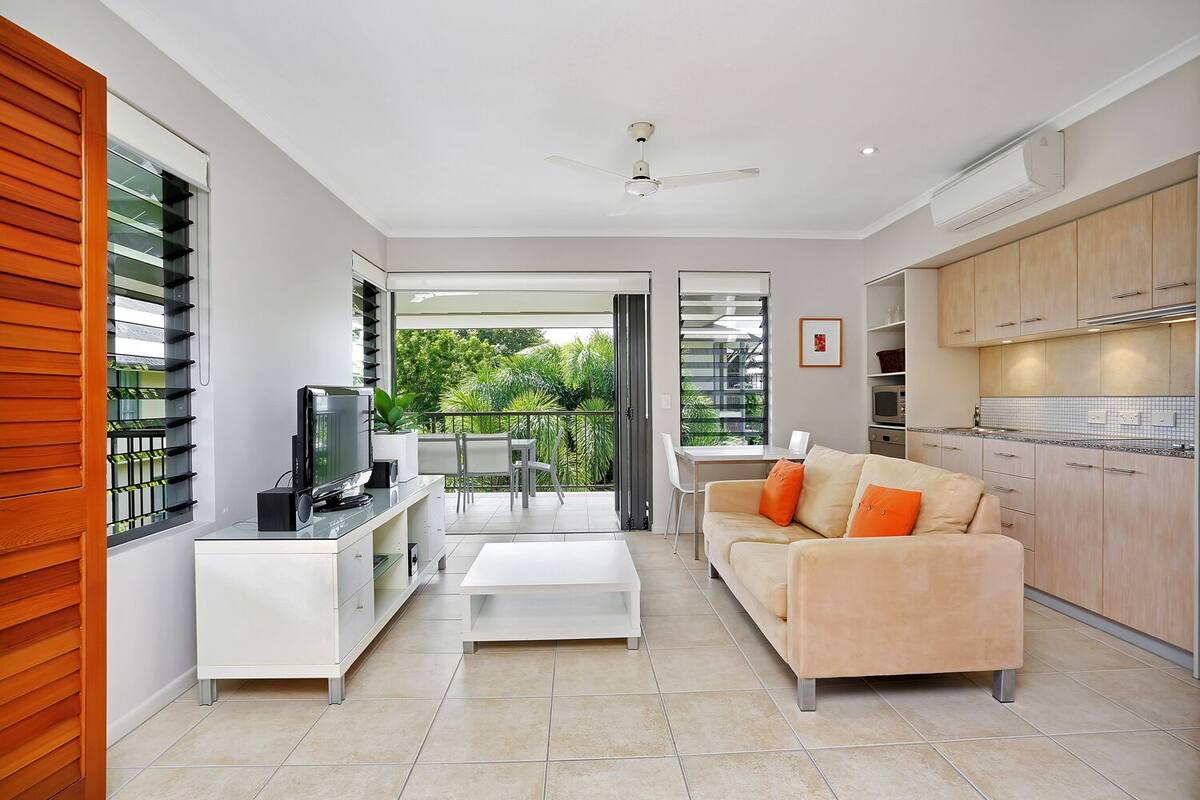 Ein modernes Wohnzimmer mit Sofa, Fernseher und Couchtisch, verbunden mit einer Küche und einem Balkon mit viel Grün.