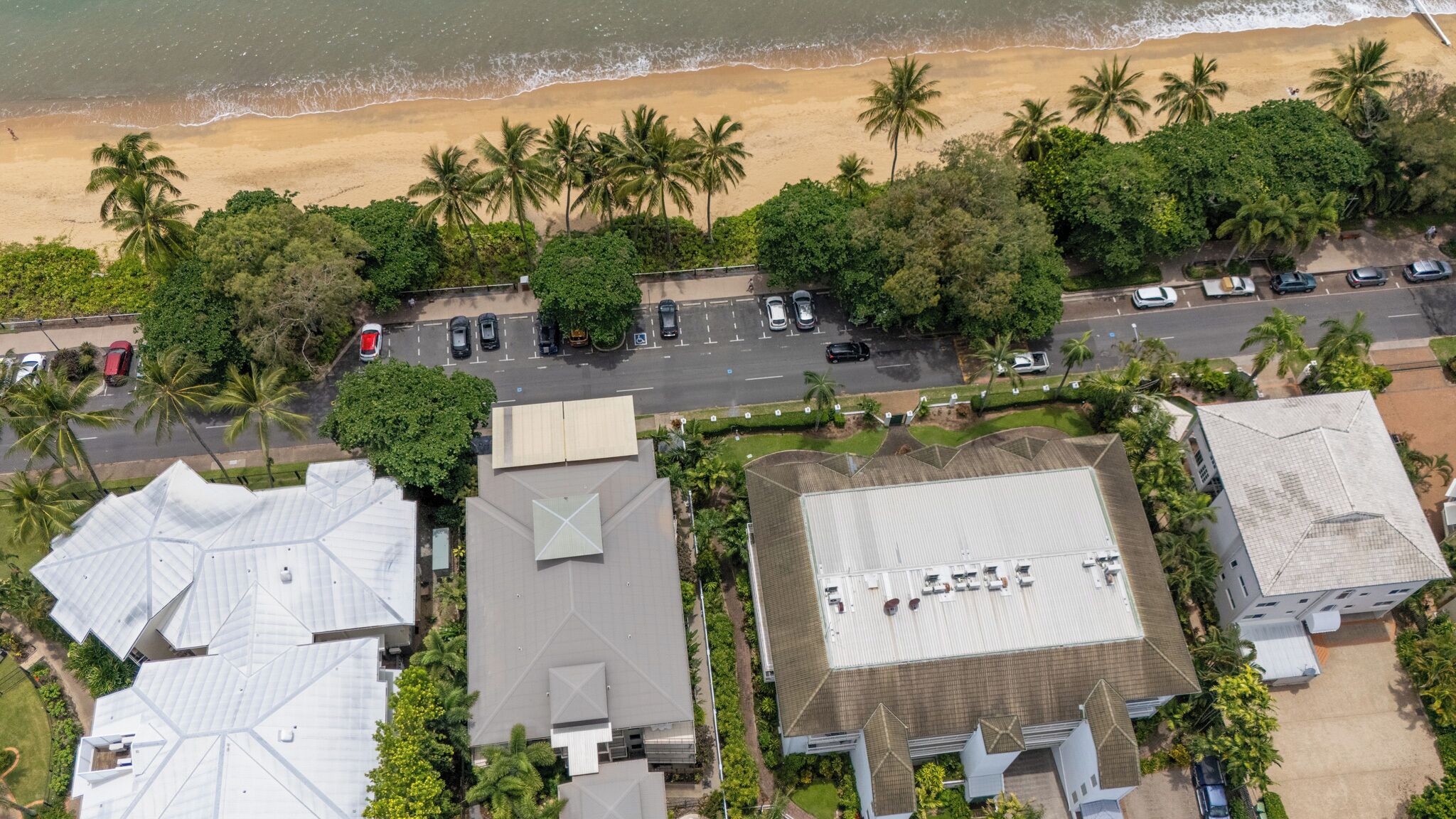 An aerial view of a coastal area with a beach, palm trees, and a parking lot with several cars.