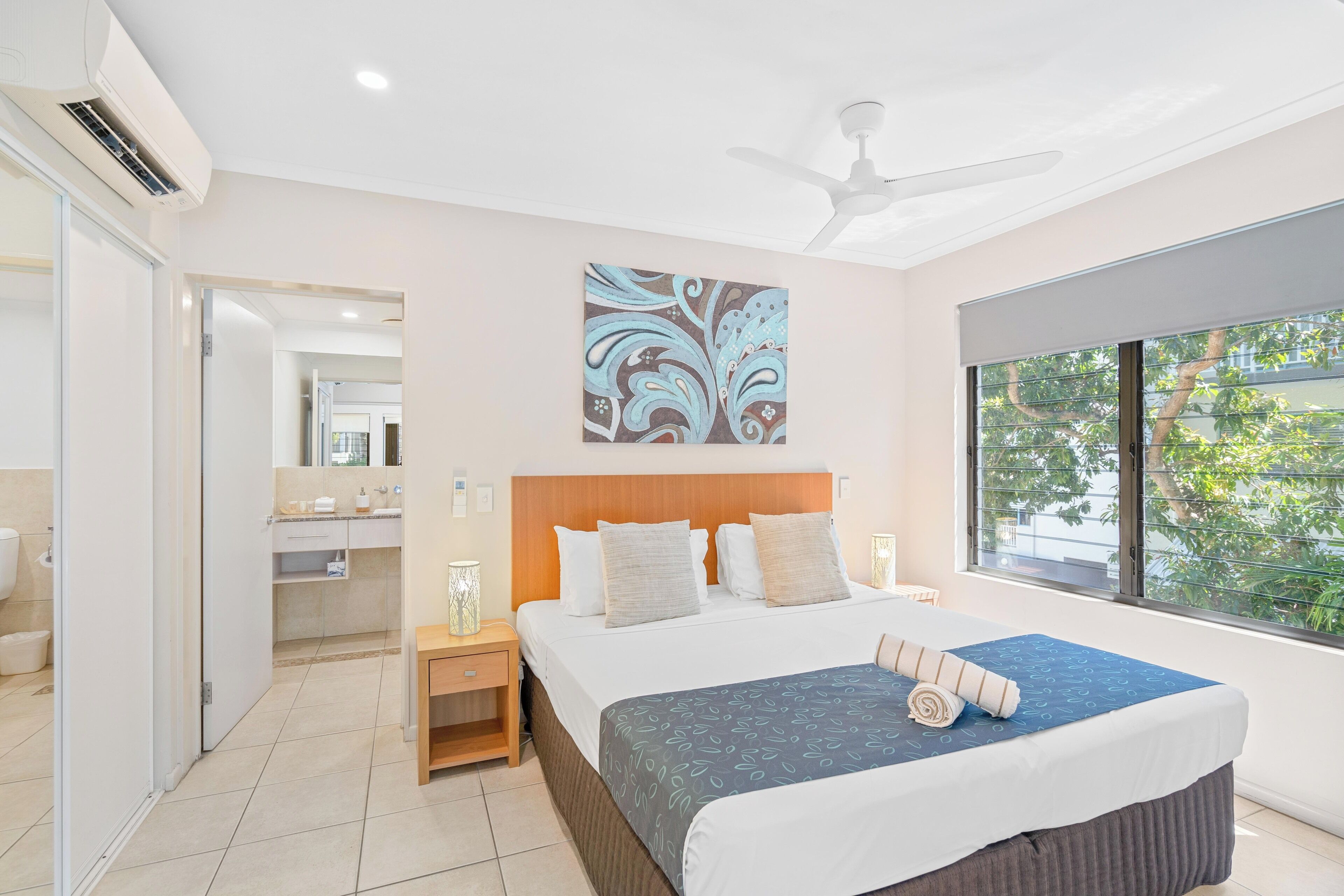 A modern hotel room with a large bed, a ceiling fan, a window with a view of greenery, and a bathroom visible through an open door.
