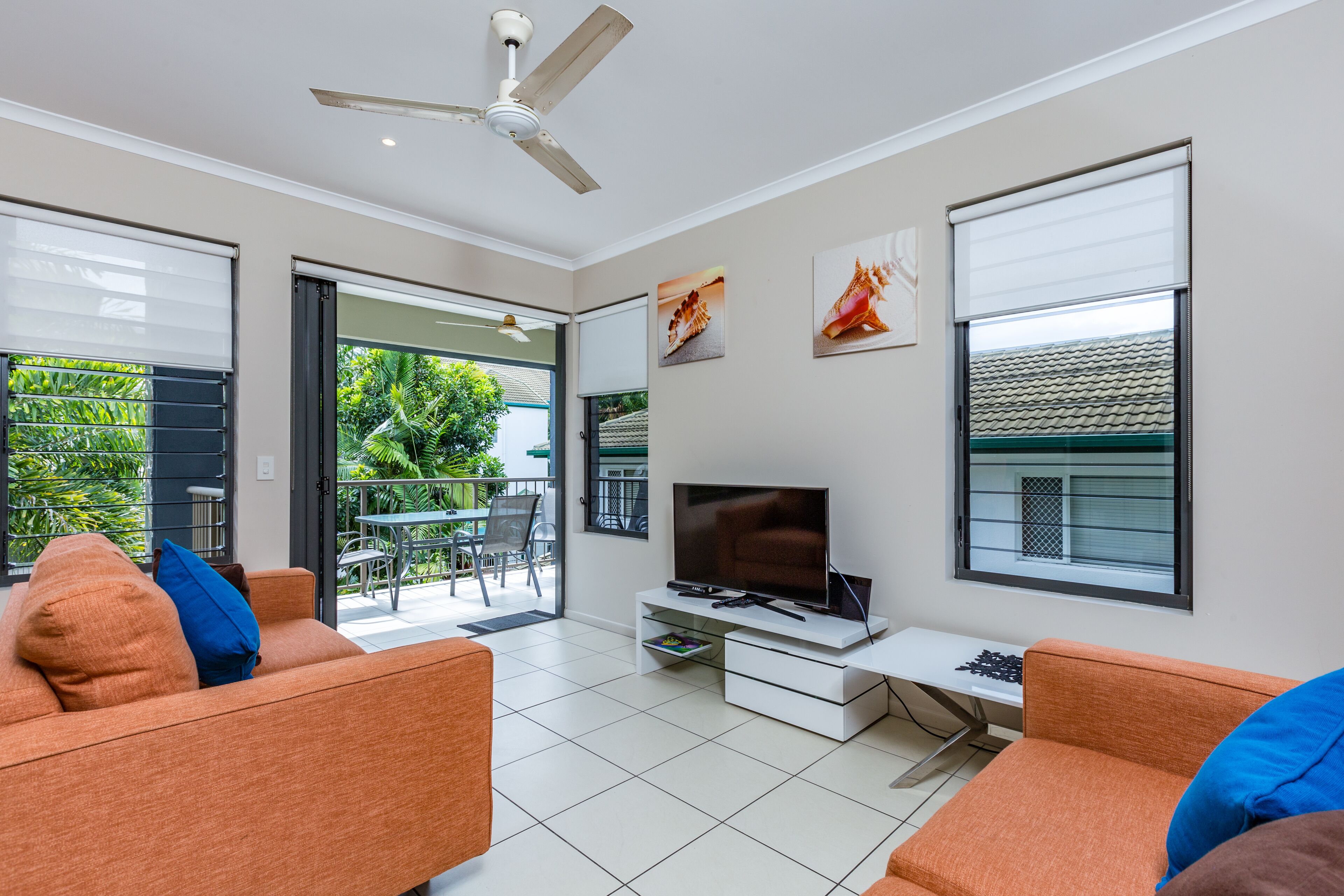 A modern living room with a flat-screen TV, two orange sofas with blue cushions, a ceiling fan, and a sliding glass door leading to a balcony with greenery.
