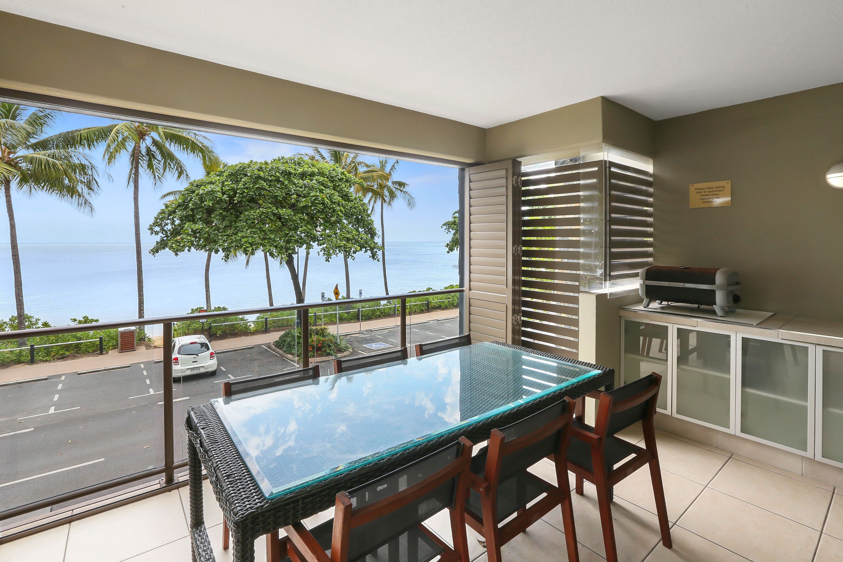 A balcony with a glass table and chairs, a view of the ocean, and a car parked outside.