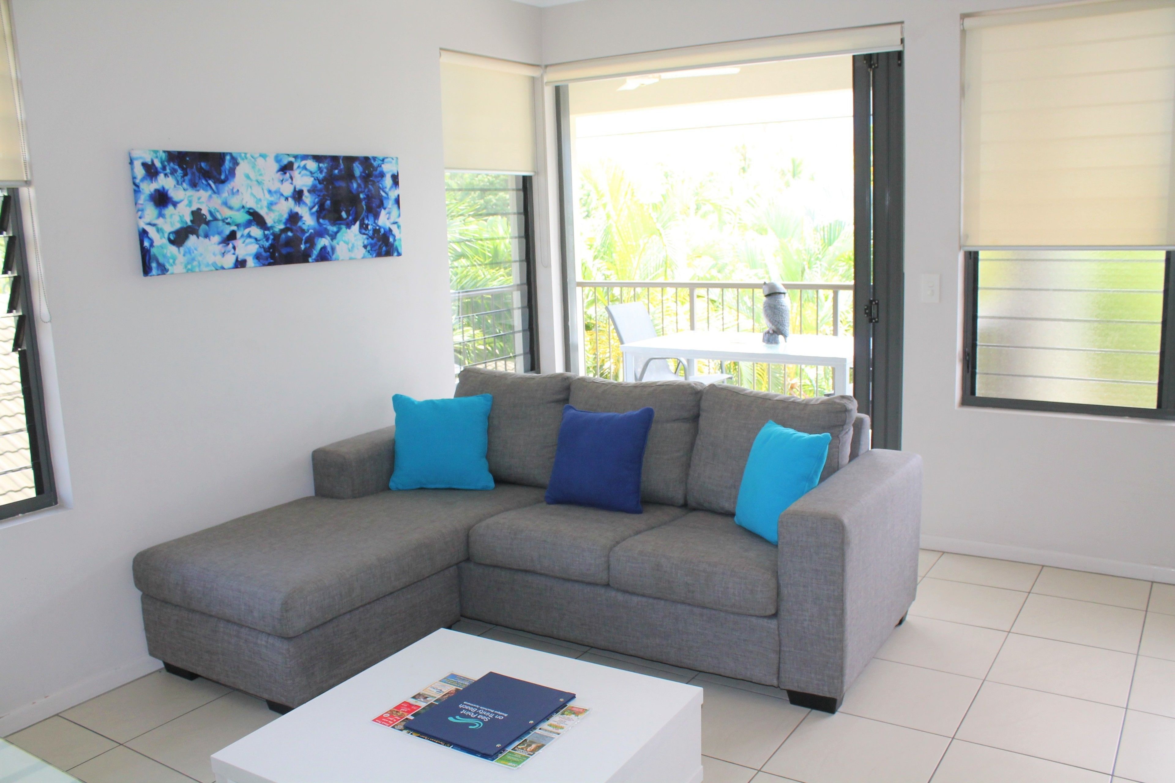 Ein Wohnzimmer mit einer grauen Eckcouch, zwei blauen Kissen, einem weißen Couchtisch mit Zeitschriften, einem großen abstrakten Gemälde und einem Balkon mit einem weißen Stuhl.