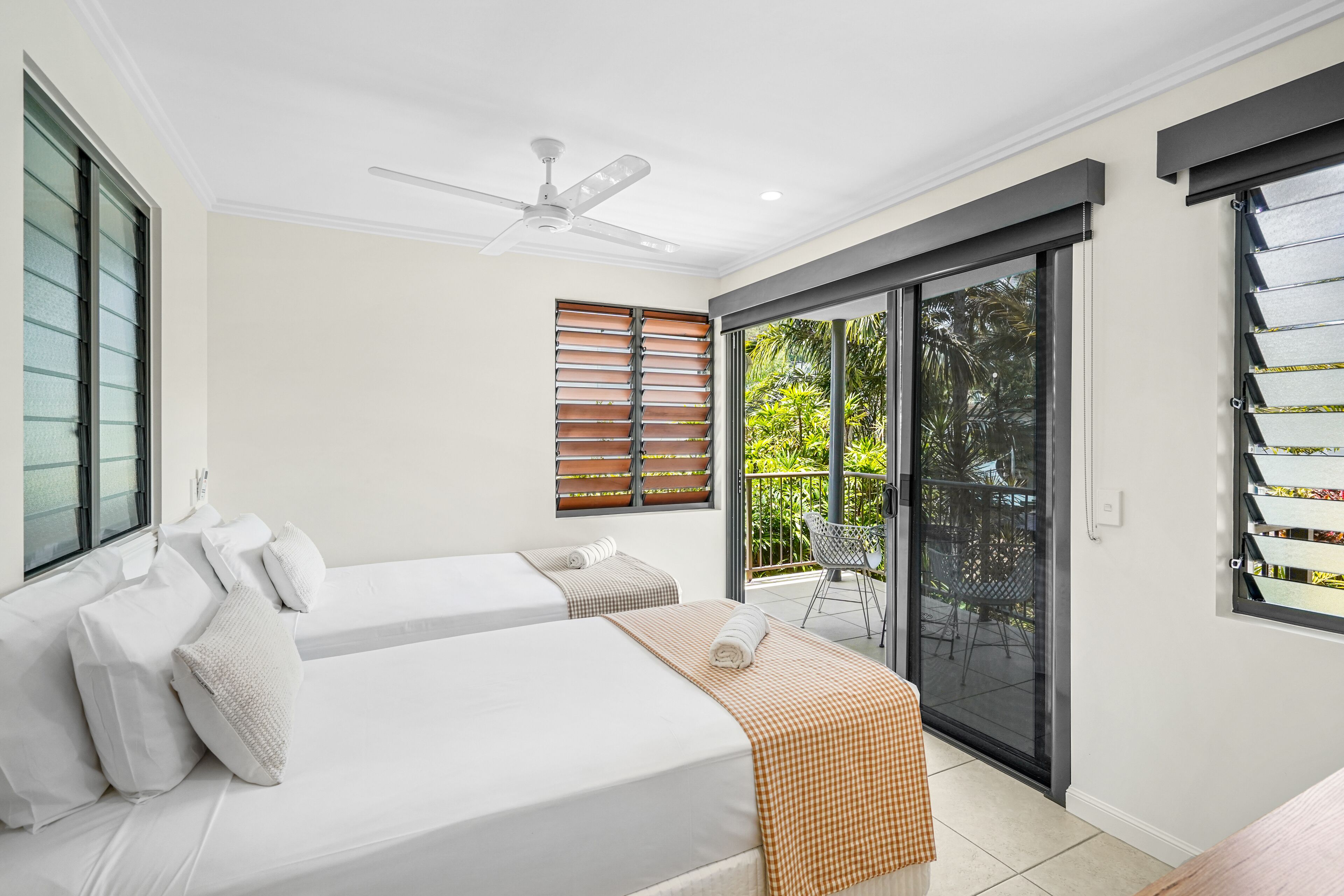 A hotel room with two beds, a ceiling fan, a sliding glass door to an outdoor area, and a view of greenery outside.