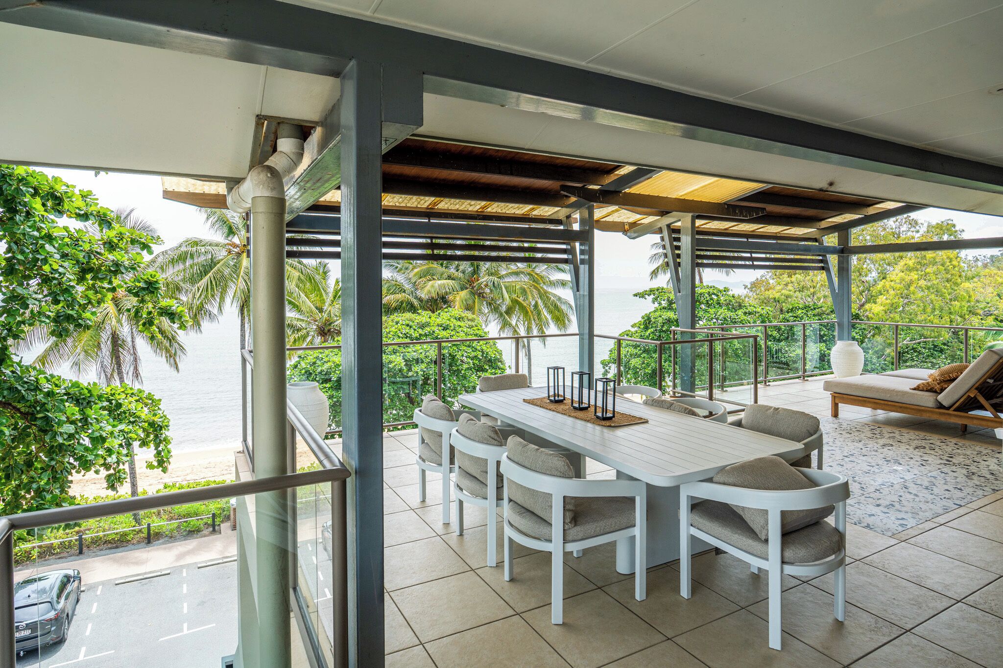 A covered outdoor dining area with a white table and chairs, overlooking a beach and palm trees.