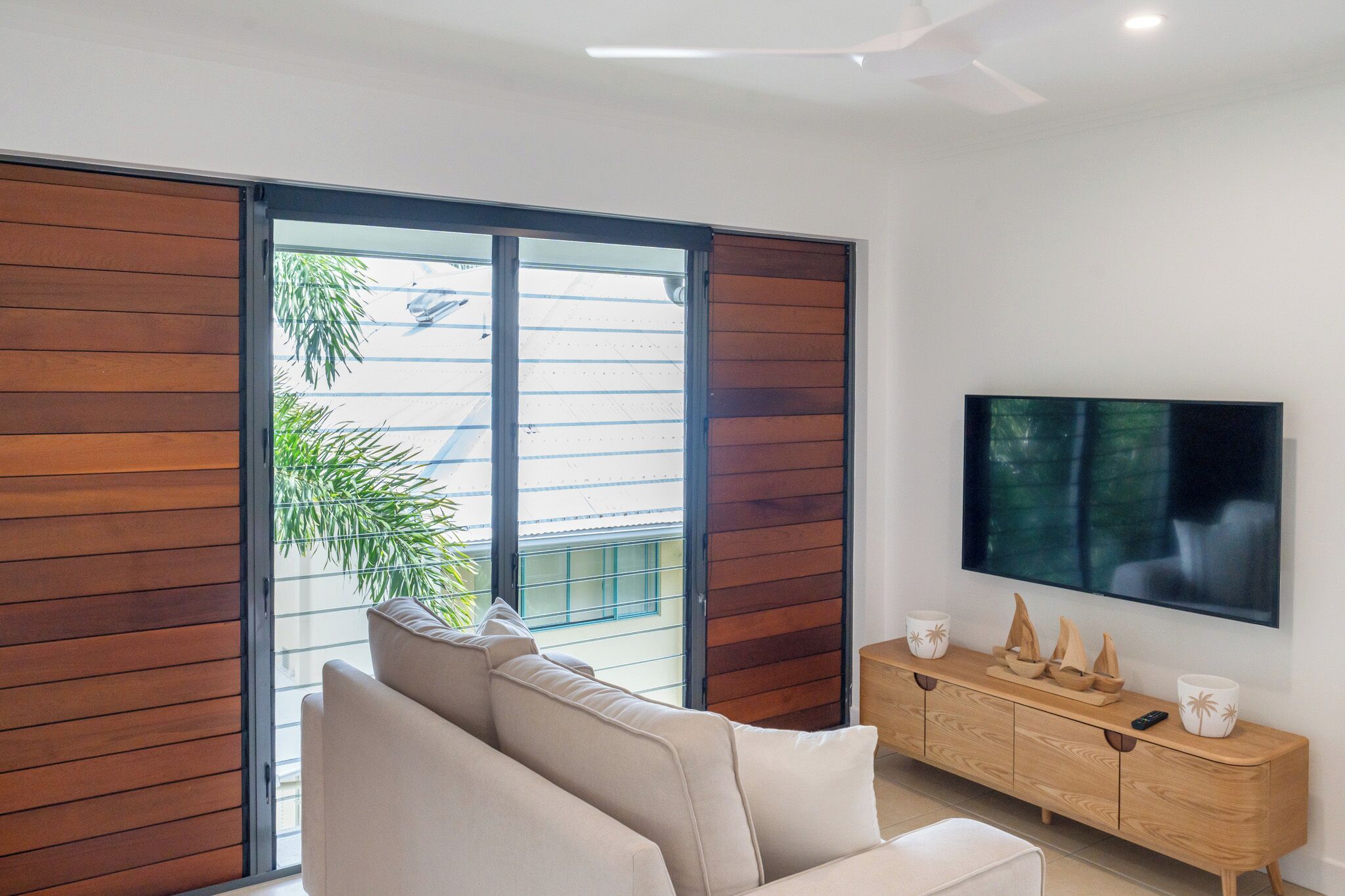 A modern living room with a sliding glass door leading to a balcony, a flat-screen TV mounted on the wall, and a wooden console table with decorative items.