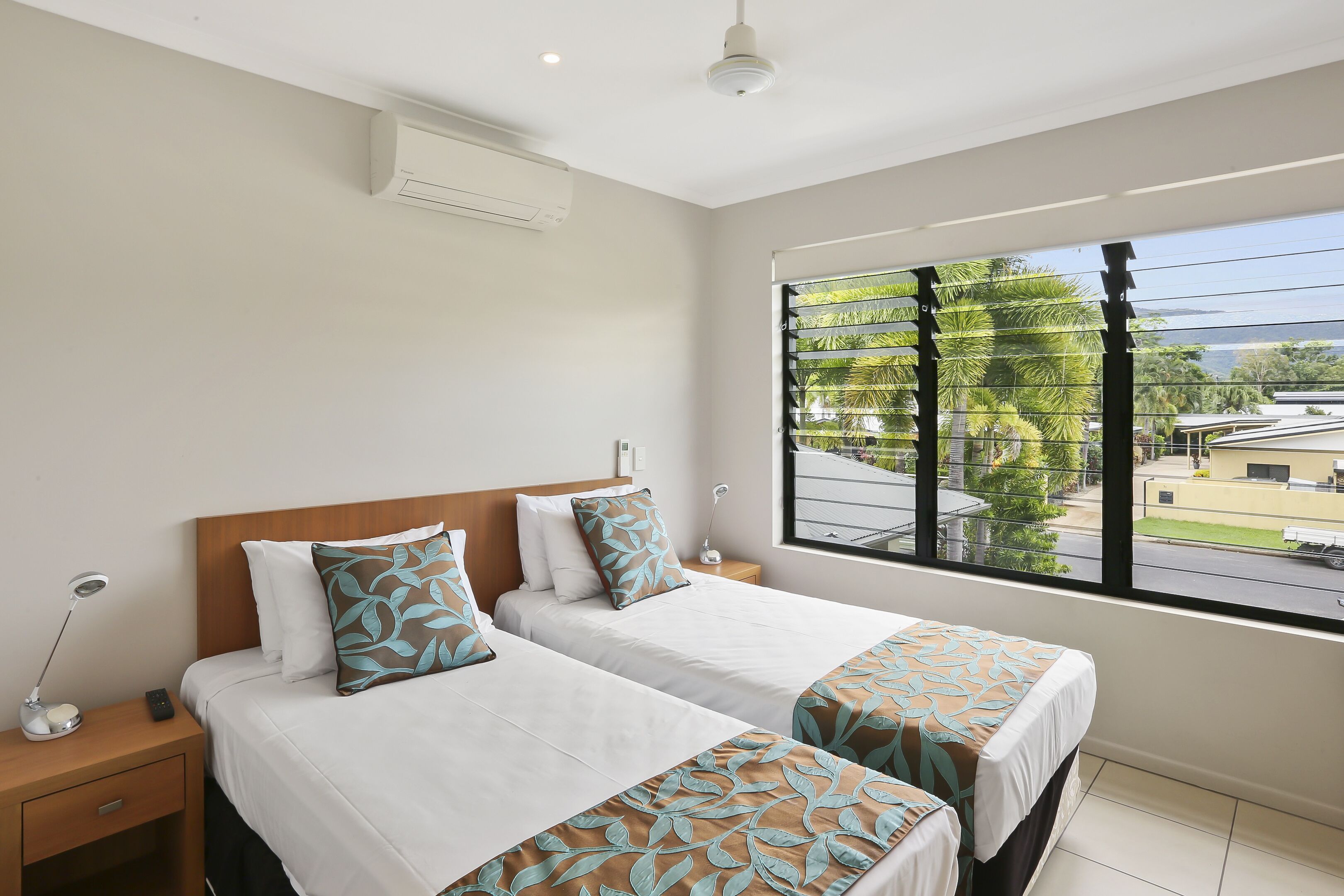A hotel room with two beds, a nightstand with a lamp, and a window with a view of palm trees and buildings.