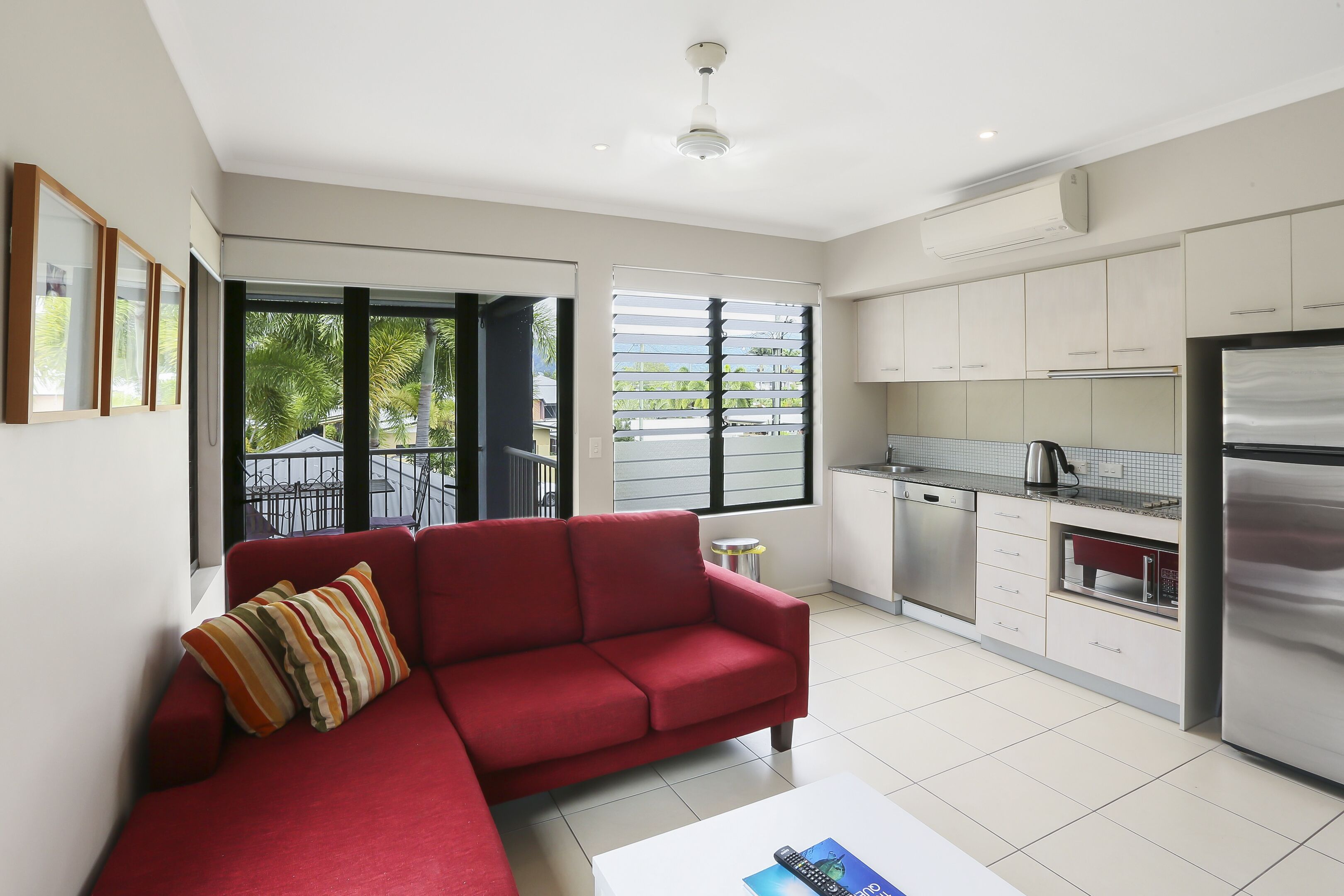 Una sala de estar moderna con un sofá rojo y una mesa de centro, una cocina con armarios y electrodomésticos blancos, y ventanas amplias con vistas al exterior.