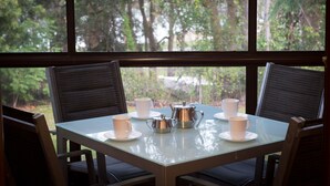 Mesa dispuesta con tazas blancas y un hervidor plateado, rodeada de sillas, con vista a árboles a través de ventanas amplias.