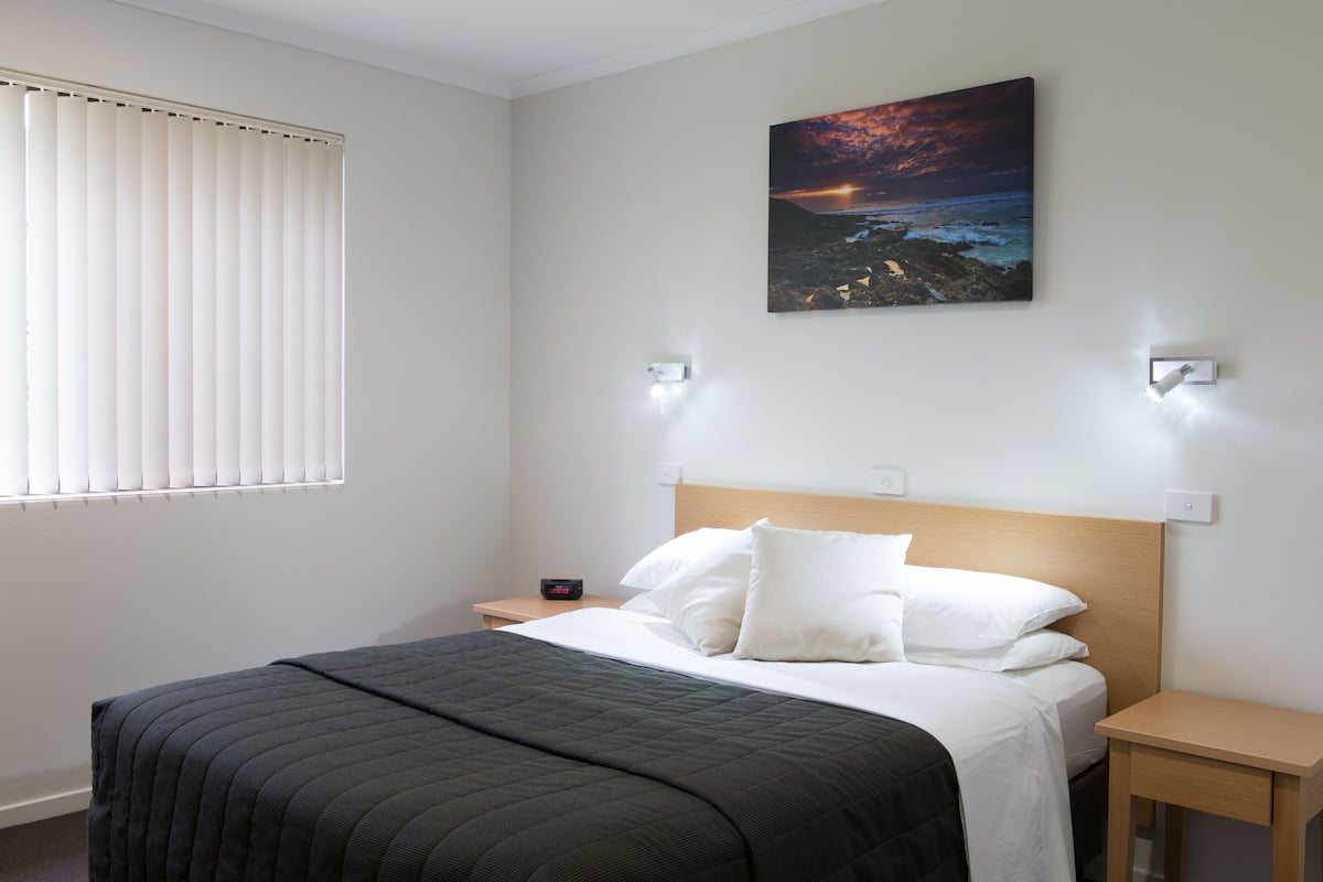 A hotel room with a bed, a nightstand, a wall-mounted painting, and a window with blinds.