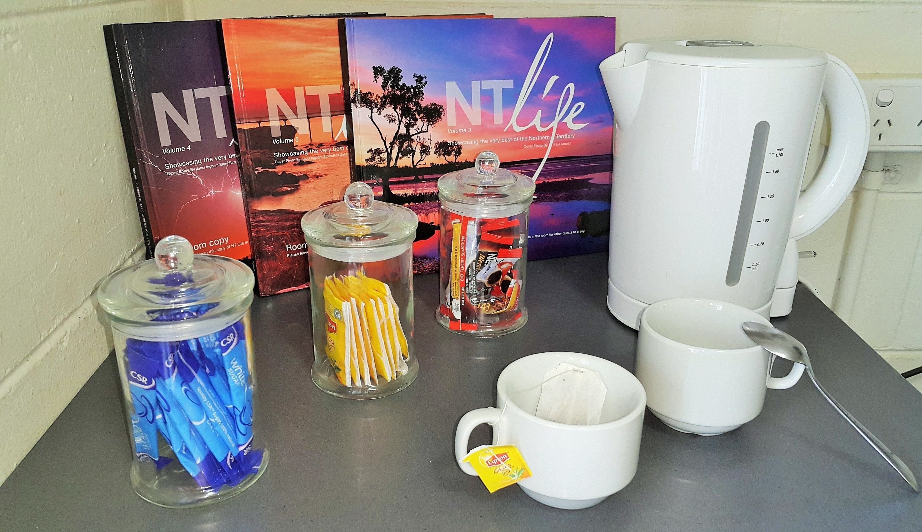 A table with a white kettle, a mug with a tea bag, and three glass containers with various items.