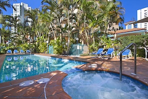 A swimming pool with a hot tub, surrounded by palm trees and blue lounge chairs. - Aruba Surf Resort (Broadbeach)