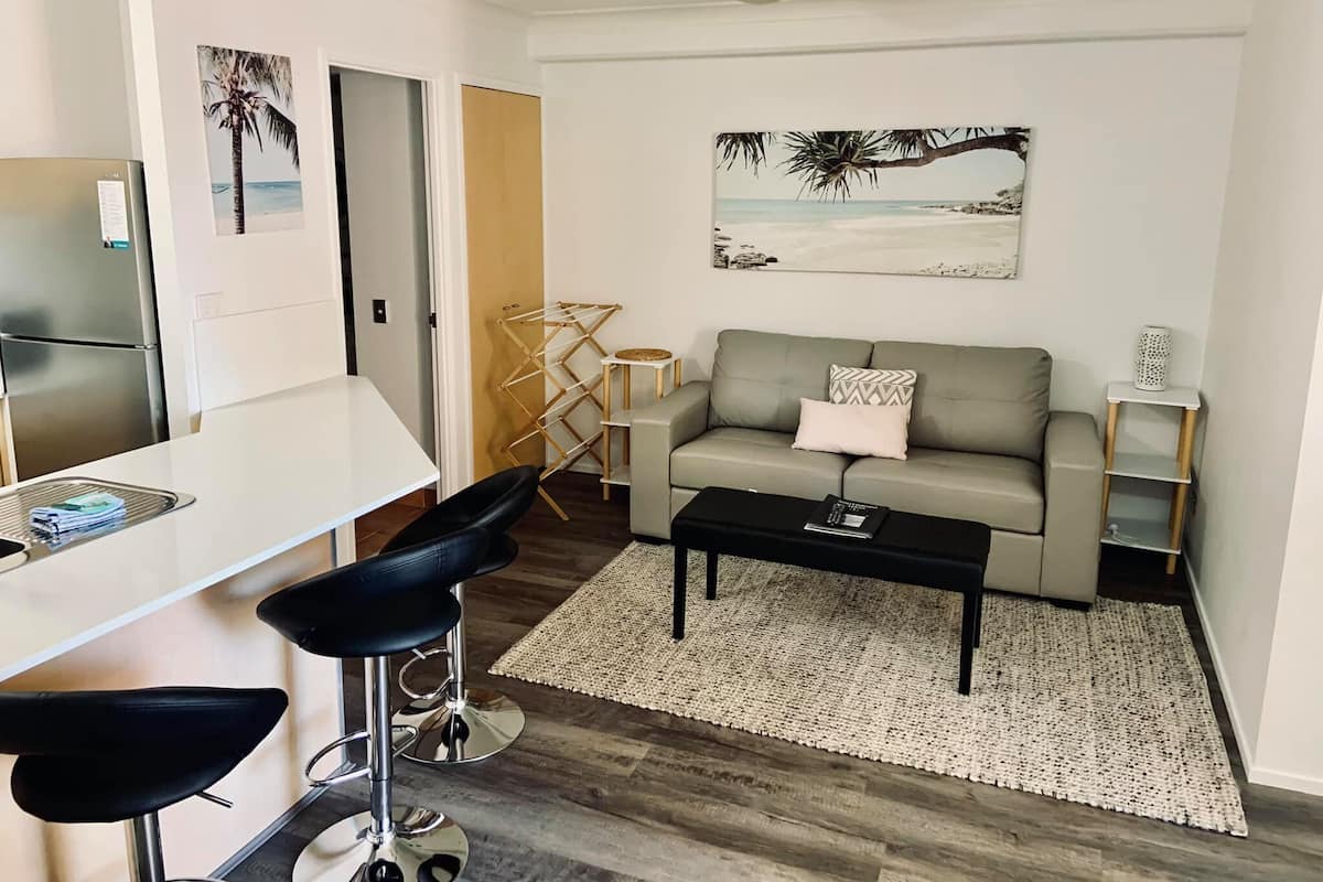 Una sala de estar moderna con un sofá gris, una mesa de centro y una zona de bar con taburetes. Hay un ventilador de techo, un cuadro de una playa y un tendedero de madera.