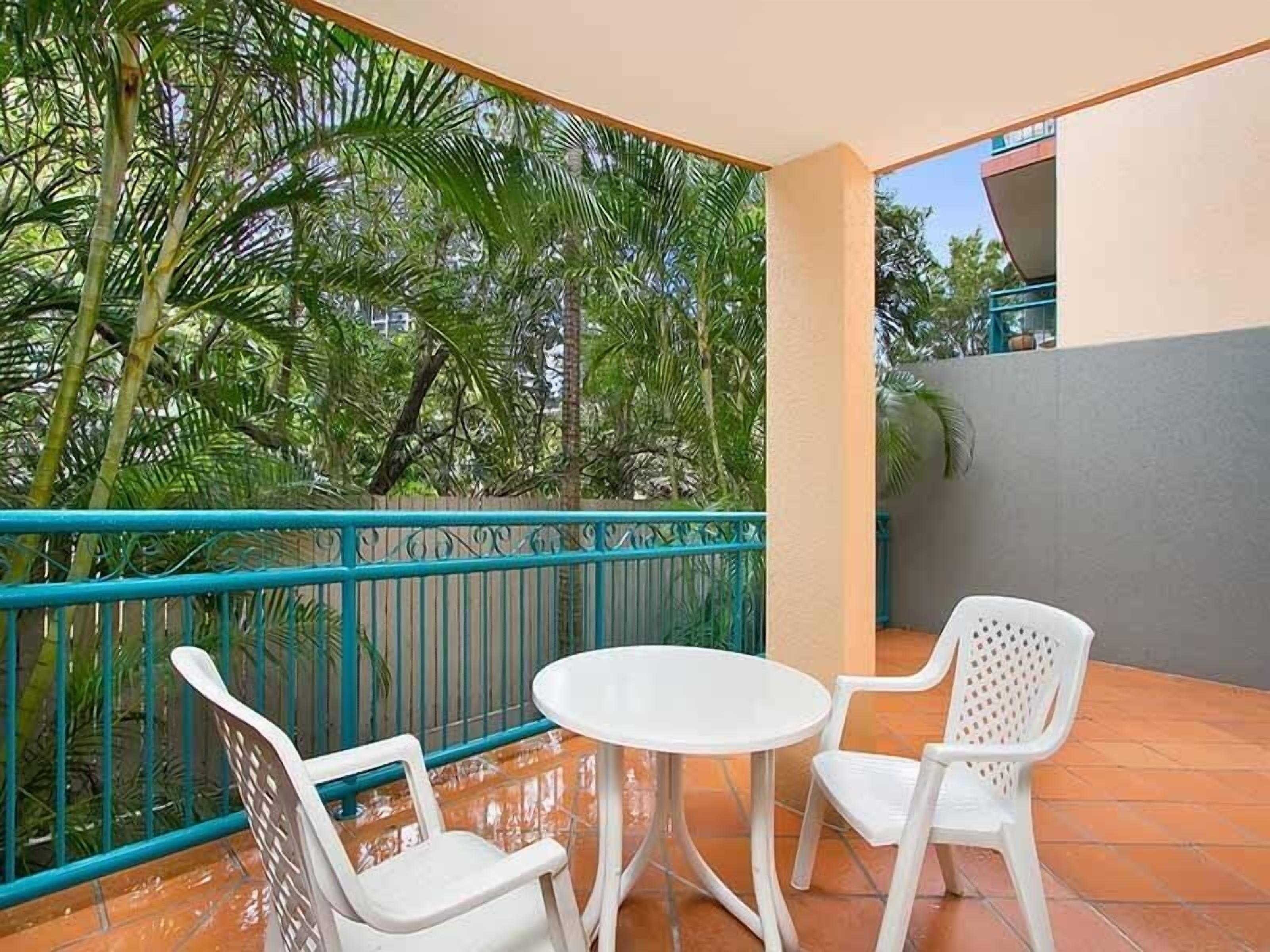 Ein Balkon mit zwei weißen Plastikstühlen und einem kleinen runden Tisch, umgeben von einem türkisblauen Geländer.