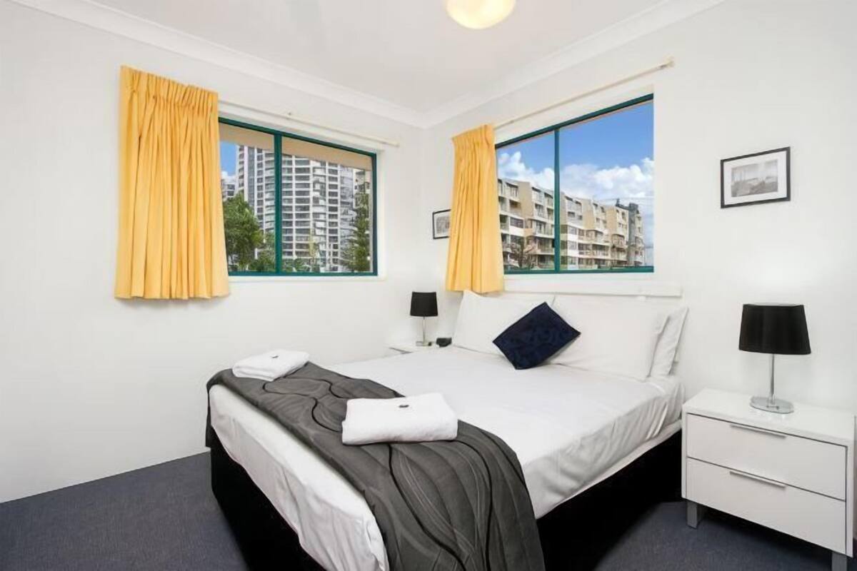 Habitación de hotel con cama, mesitas de noche, ventana con cortinas, un cuadro en la pared y vistas a edificios.