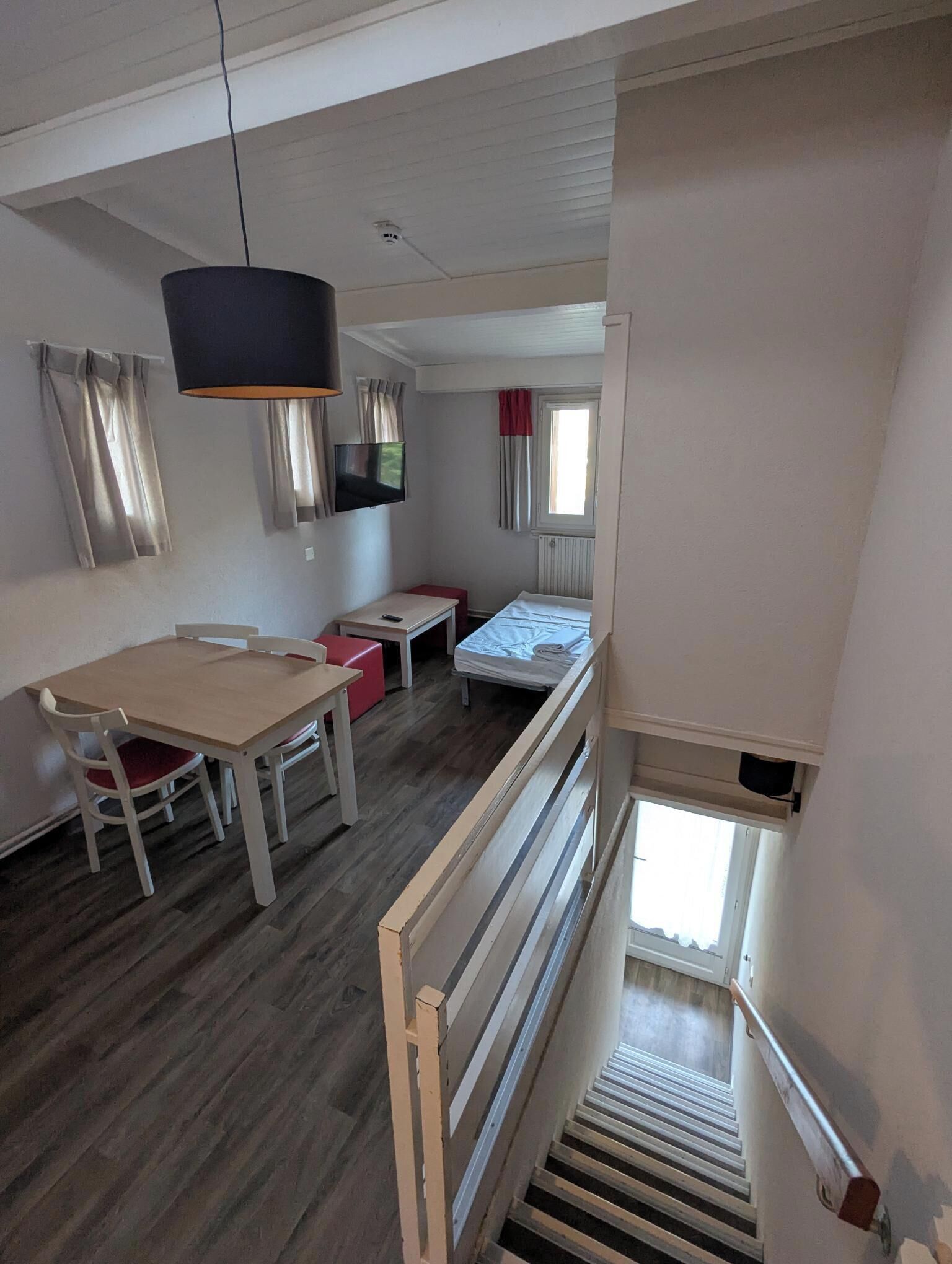 Una habitación con piso de madera, una cama, un escritorio, una silla, un televisor y una ventana con cortinas.