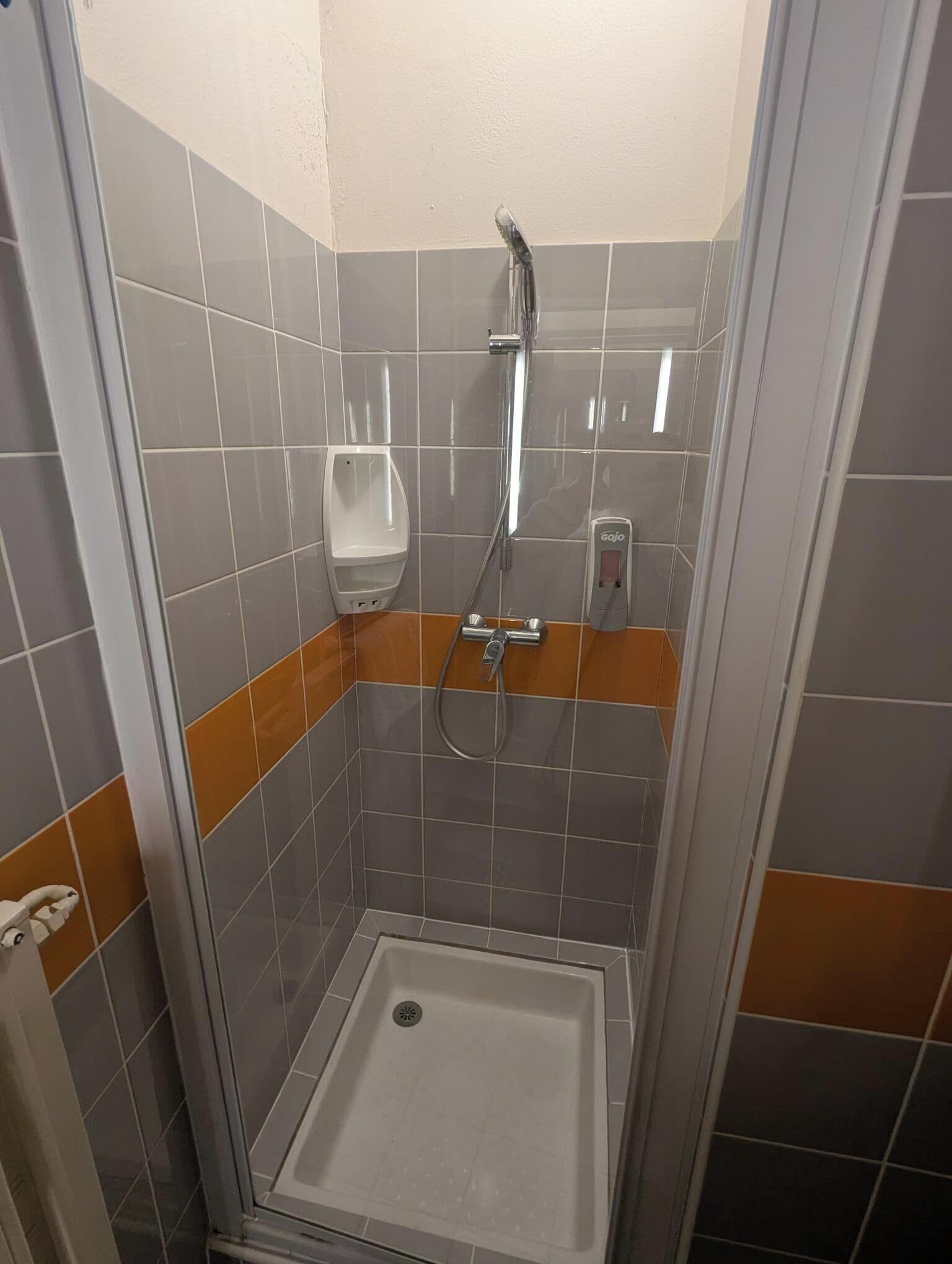 Eine Duschkabine mit Glastür, einer Handbrause, einem an der Wand befestigten Seifenspender und einem Handseifenspender.