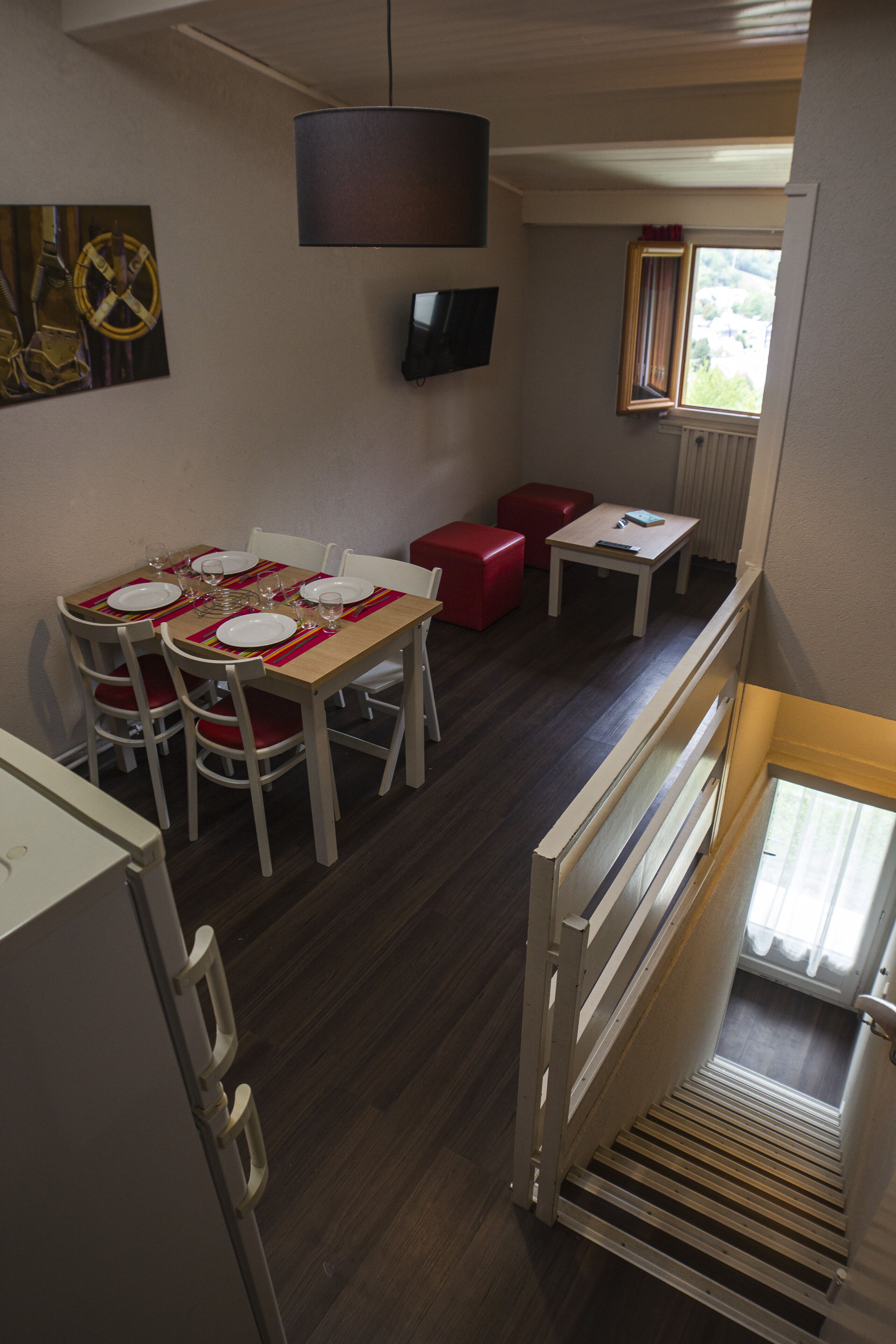 Un área de comedor con una mesa dispuesta para cuatro, un televisor montado en la pared y una ventana con cortinas.