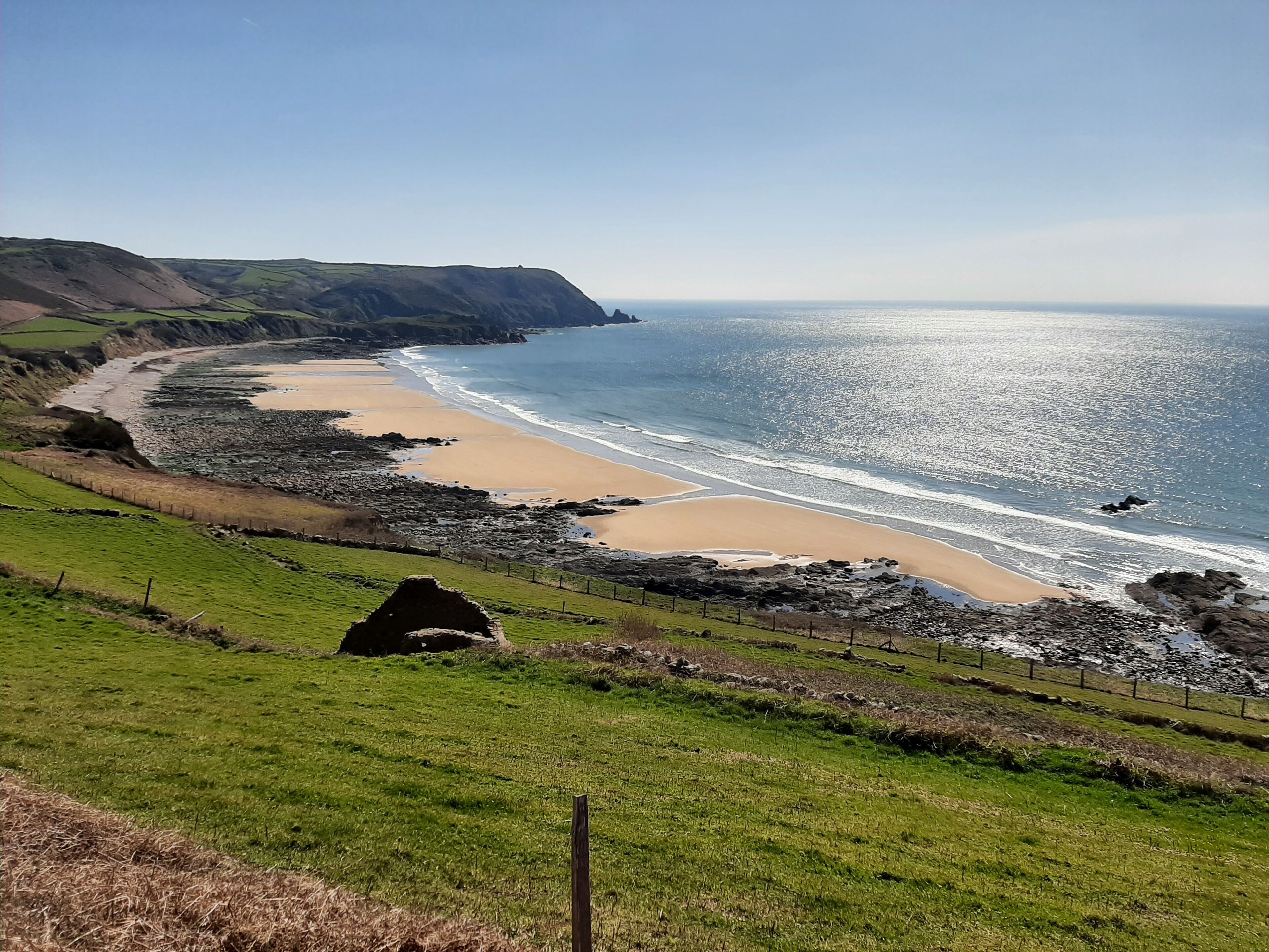 Un paisaje costero con una playa de arena, un litoral rocoso y campos cubiertos de hierba.