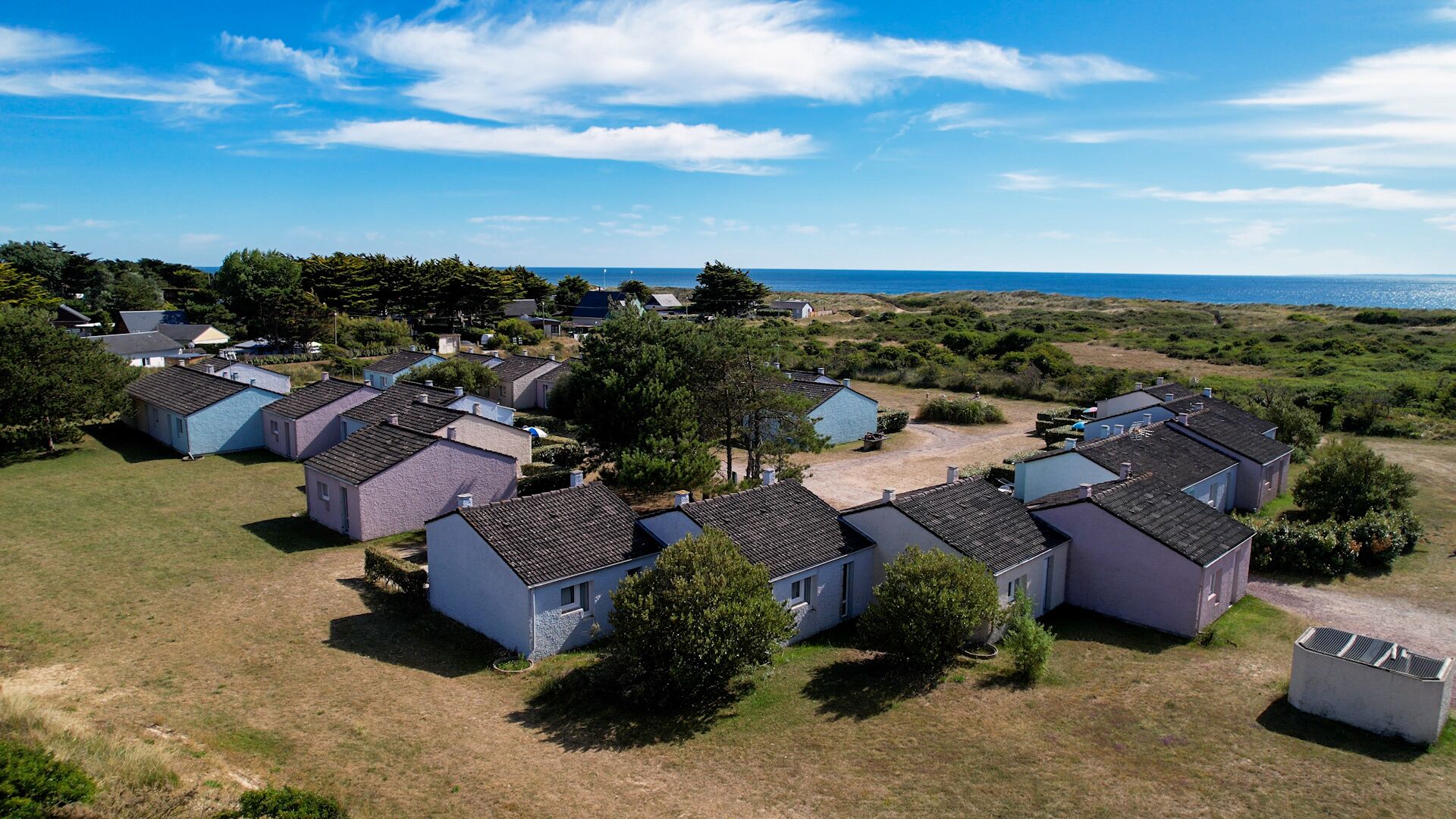 A cluster of small, single-story houses with sloped roofs, arranged in a semi-circular pattern around a central grassy area, with a view of the sea in the background.