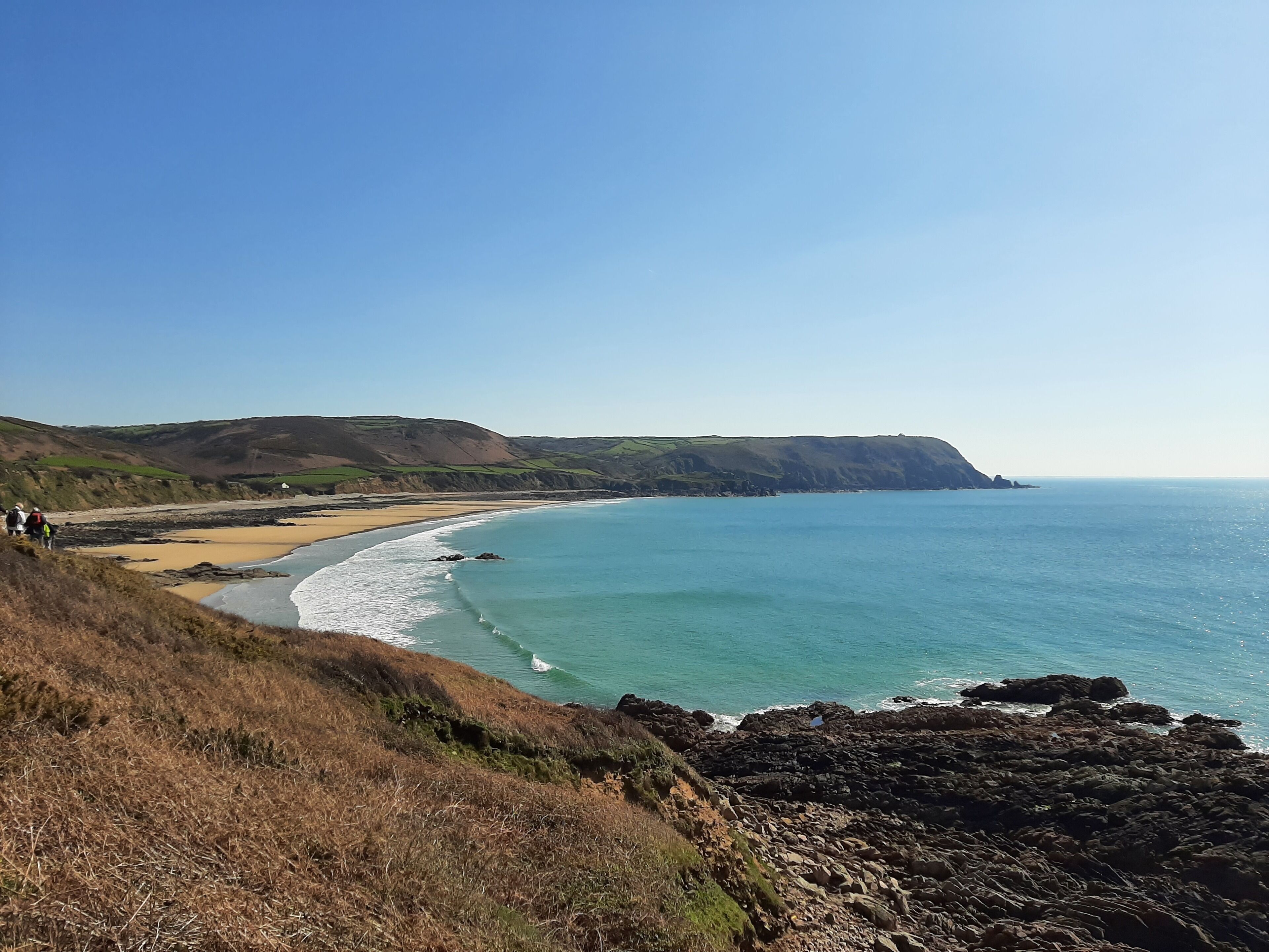 A coastal landscape with a beach, rocky shoreline, and hills in the background.