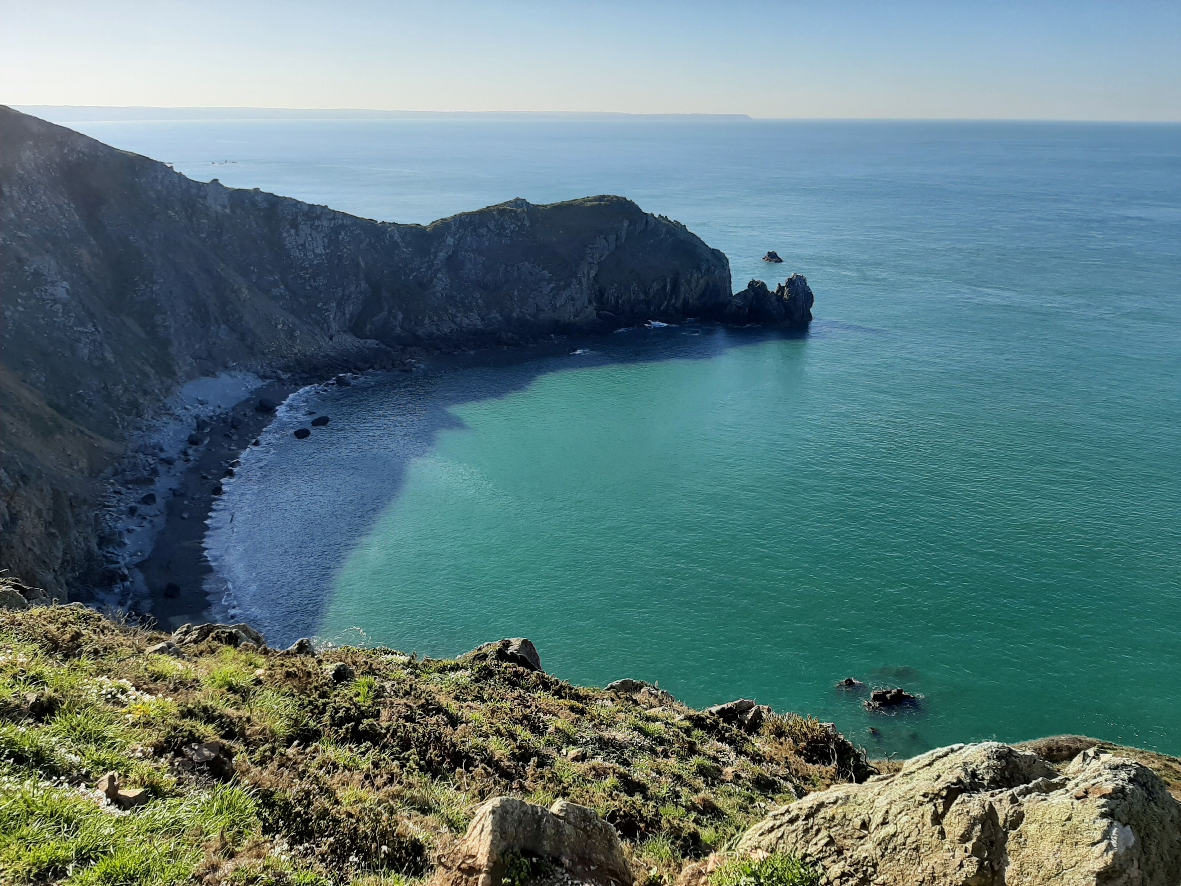 Un paisaje costero con un acantilado rocoso, un mar turquesa transparente y un pequeño promontorio pedregoso.