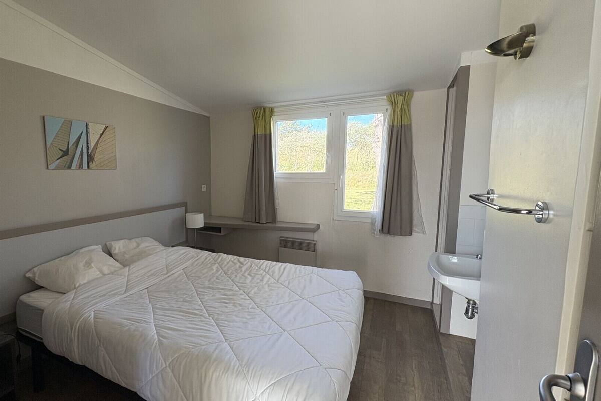 A hotel room with a bed, bedside table, lamp, window with curtains, and a bathroom with a sink and mirror.