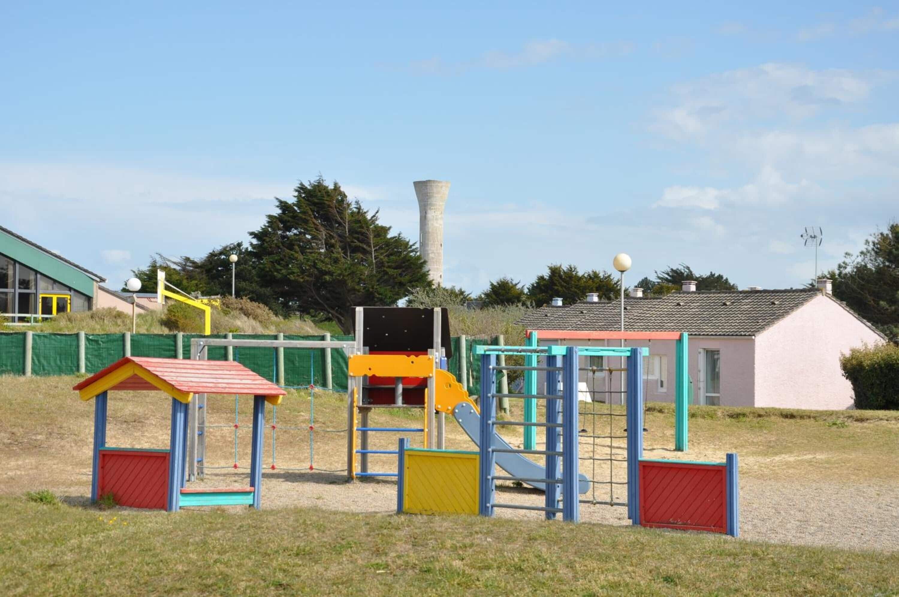 A playground with colorful play structures, a tall chimney, and a building with a red roof in the background.