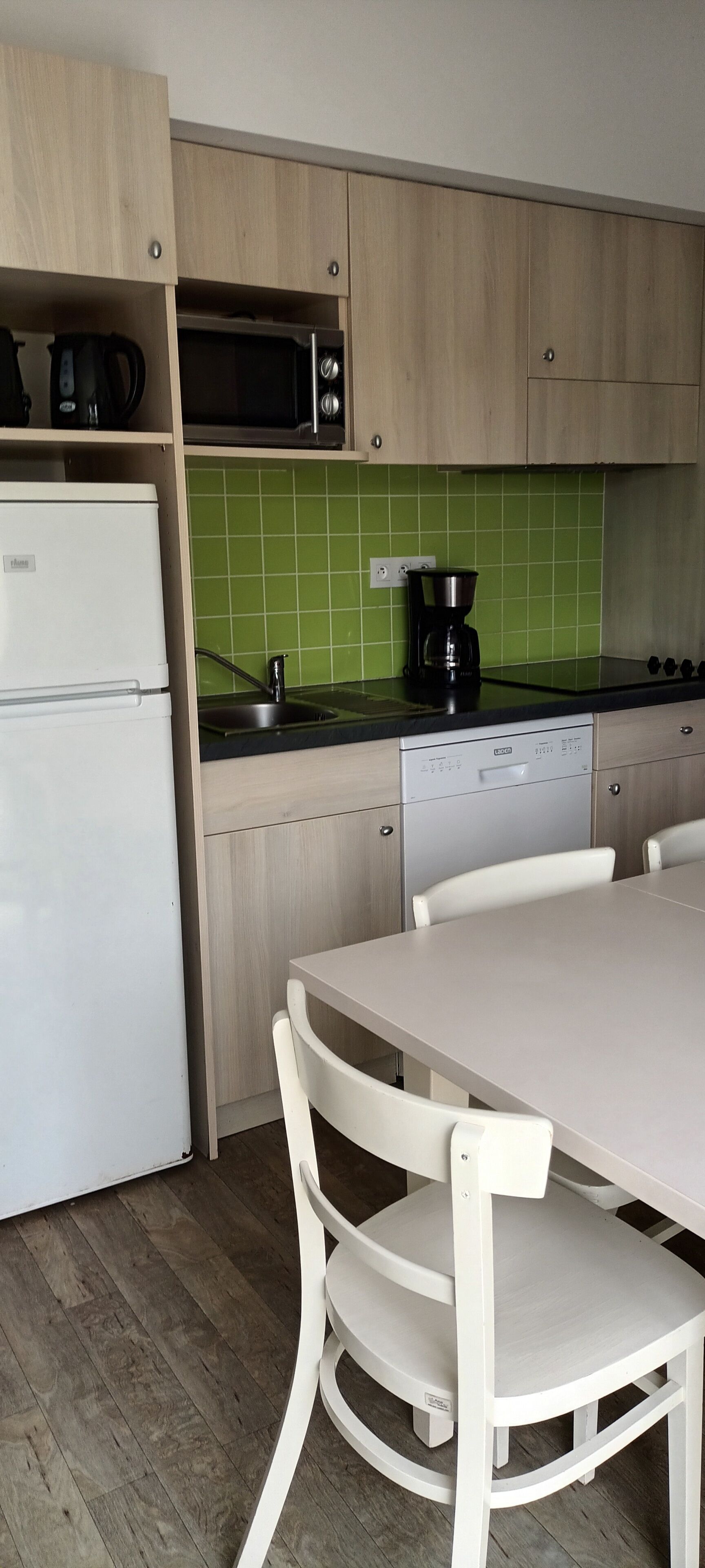 Uma cozinha compacta com armários de madeira, uma geladeira branca, micro-ondas, cafeteira e uma mesa de jantar com cadeiras.